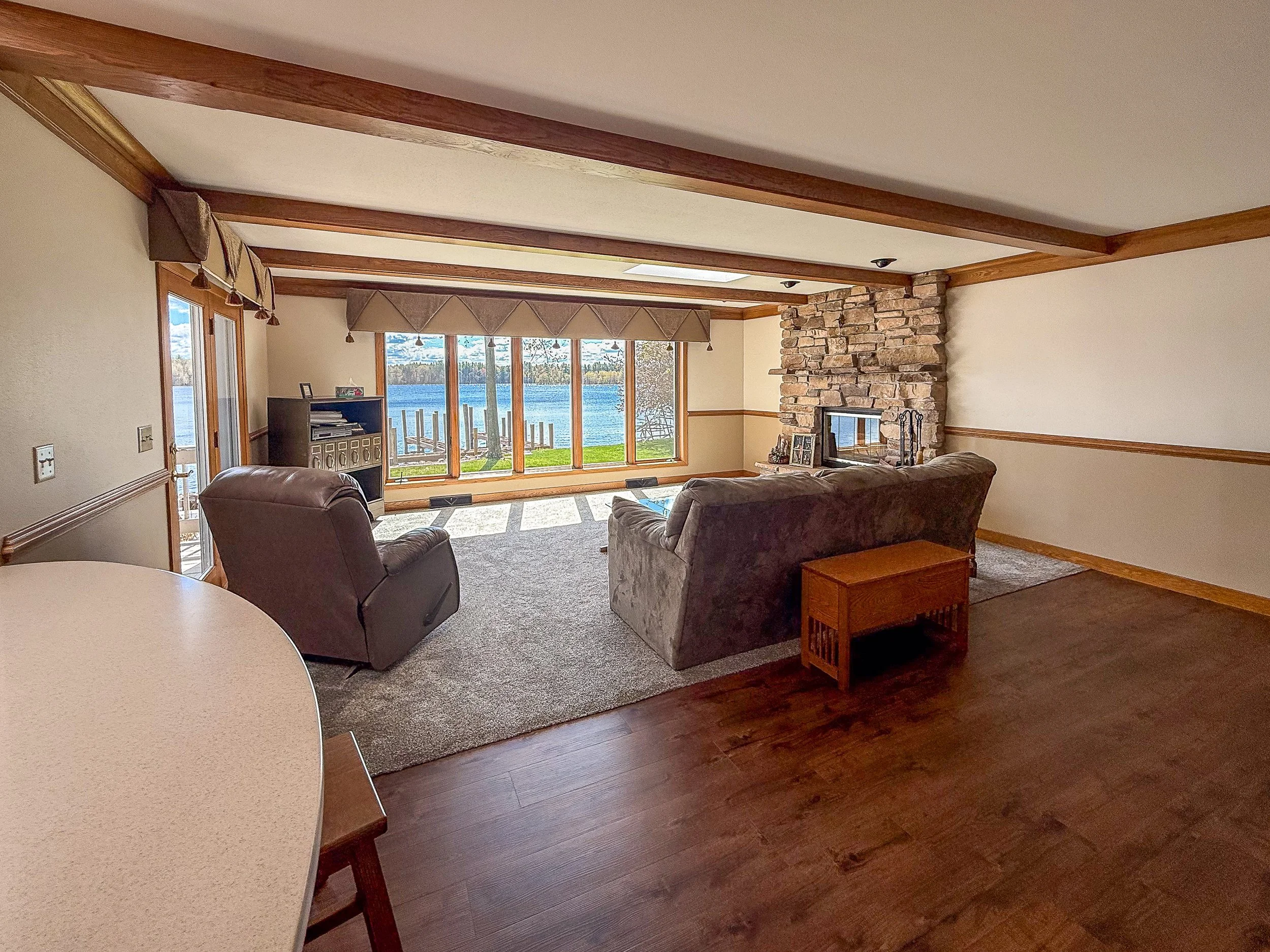 Living room with large window overlooking a lake, stone fireplace, brown couches, wooden flooring, and a window valance with tassels.