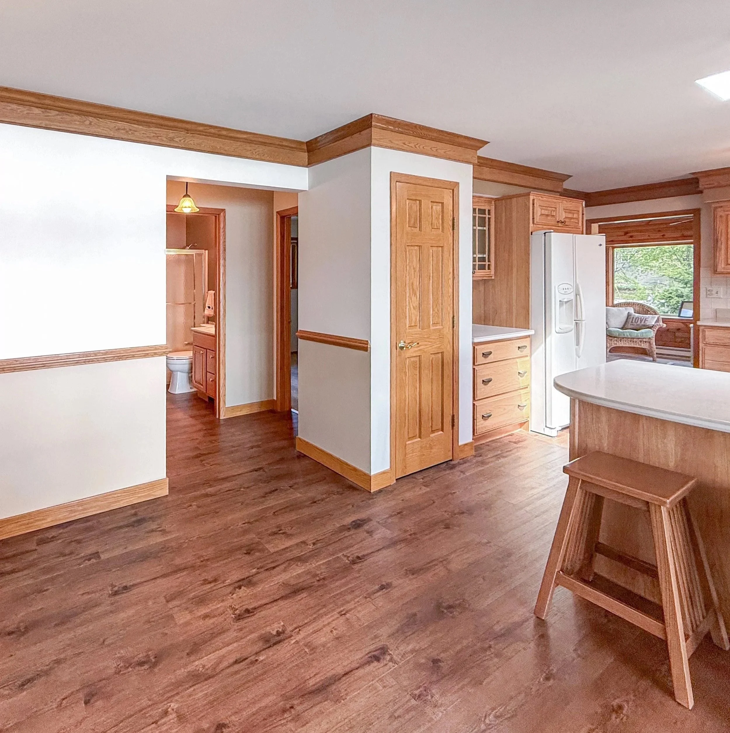 Living room with hardwood floors, wooden crown molding, and a wooden dining table with chairs. A white refrigerator is visible near a window with a garden outside. Part of a kitchen counter and a small side room with a toilet are also seen.