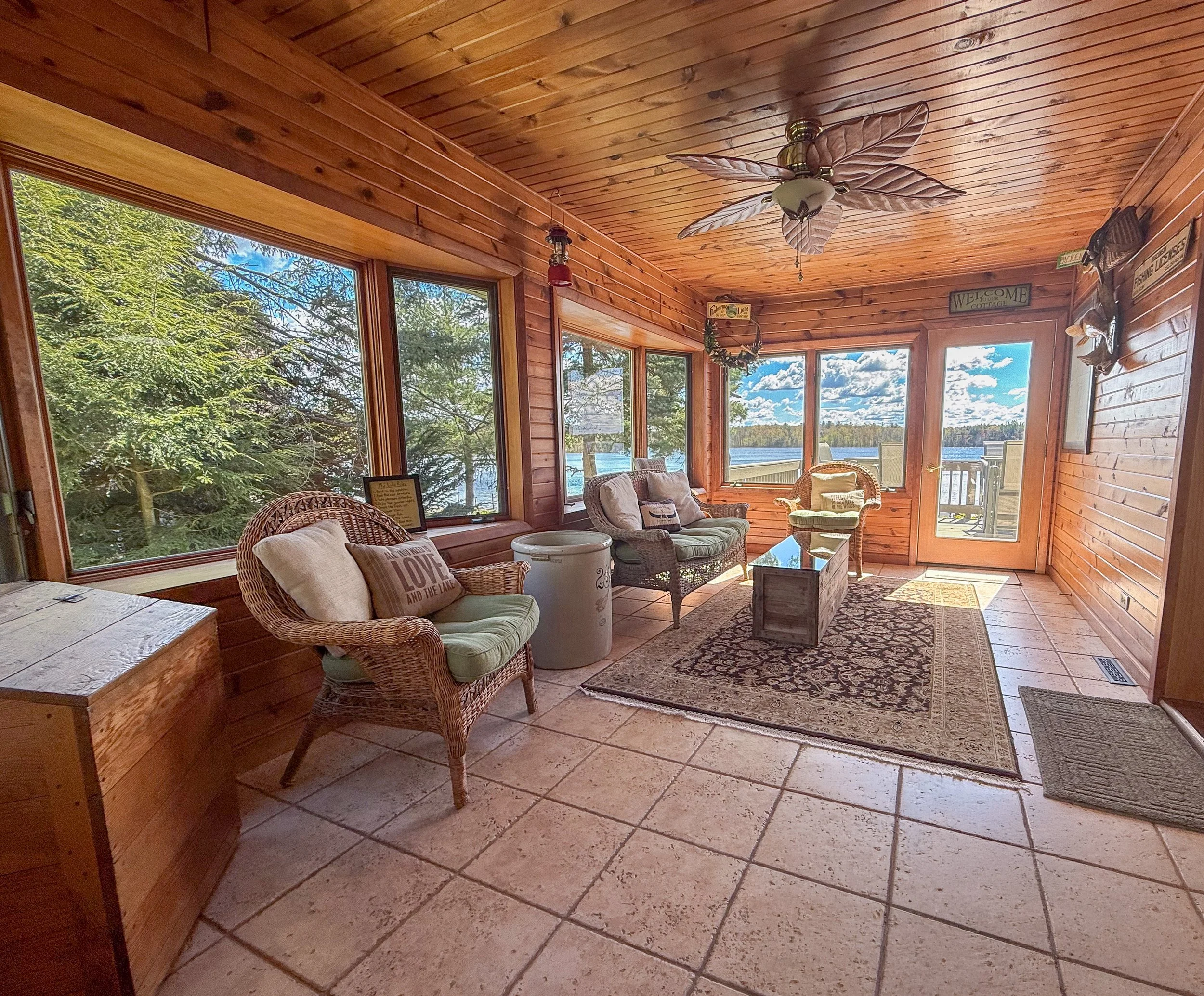 A sunroom with wood paneling, large windows overlooking a lake and trees, wicker chairs, a sofa, a coffee table, and decorative signs on the walls.