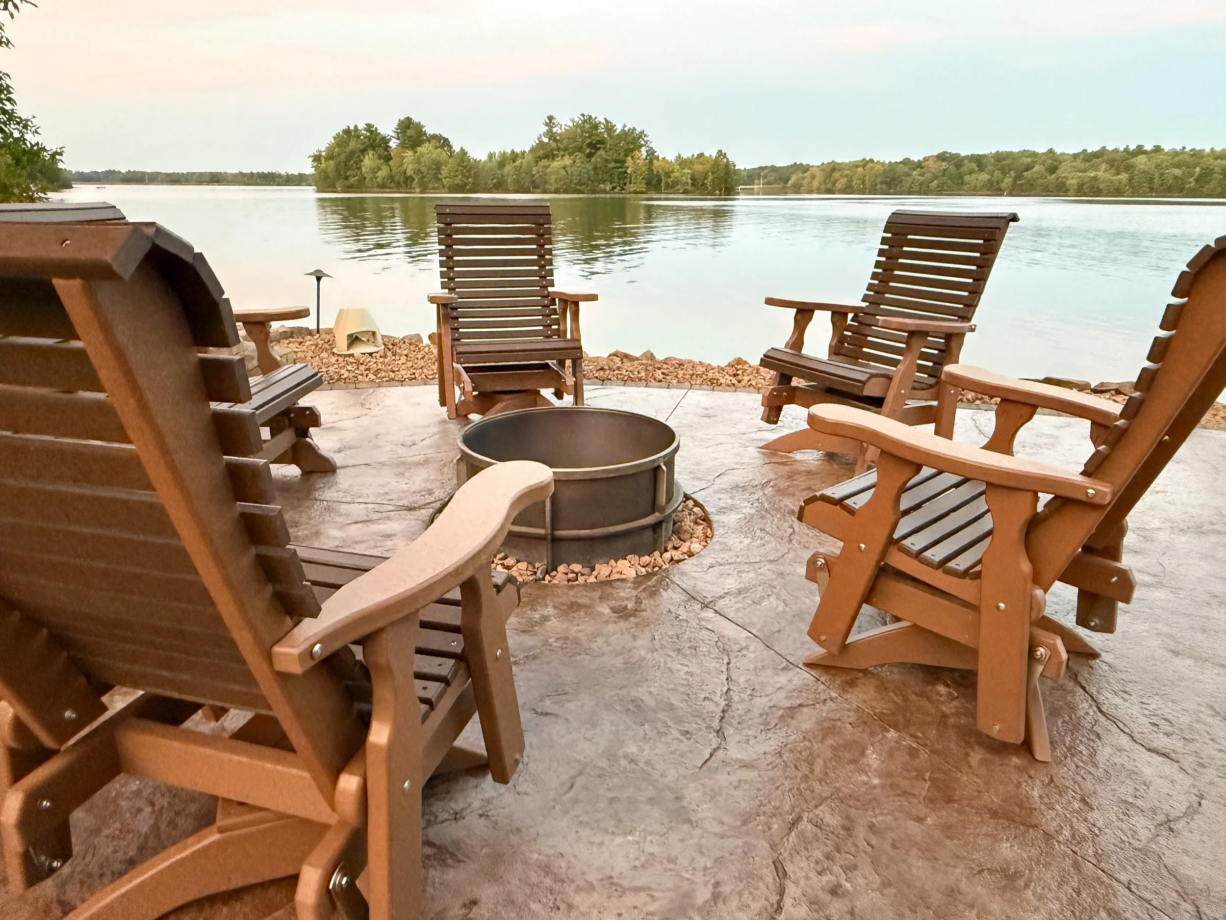 View of a lake with a wooded shoreline in the distance, seen from a patio with multiple wooden chairs and a fire pit in the foreground.