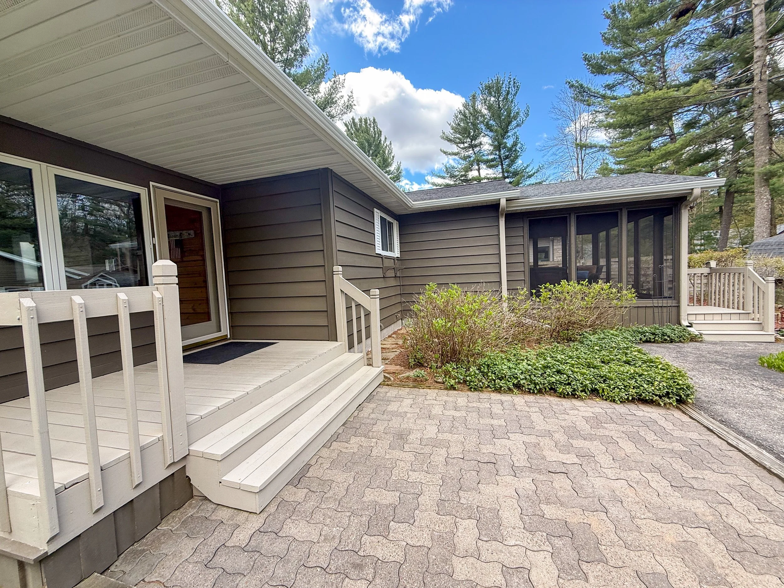 Back porch with wooden stairs and black metal railing, beige siding, sliding glass door, screened-in porch, brick paved ground, garden bushes, tall pine trees in background under partly cloudy sky.