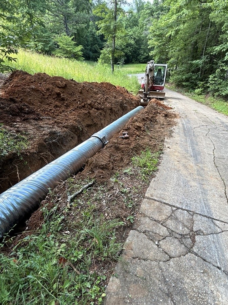 A large pipe runs through a trench beside a cracked paved road, with a small excavator nearby and green trees in the background.