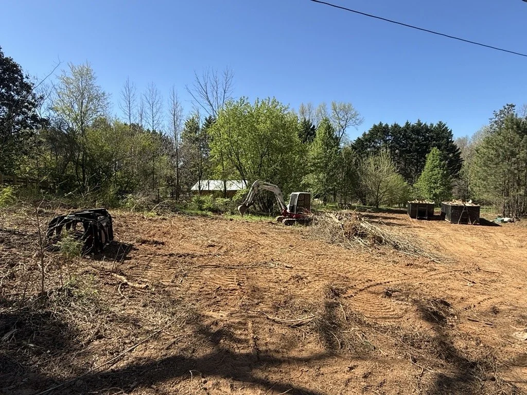 Cleared land with dirt, small tree branches, a small excavator, and several trash bins, with trees and a house in the background under a clear blue sky.