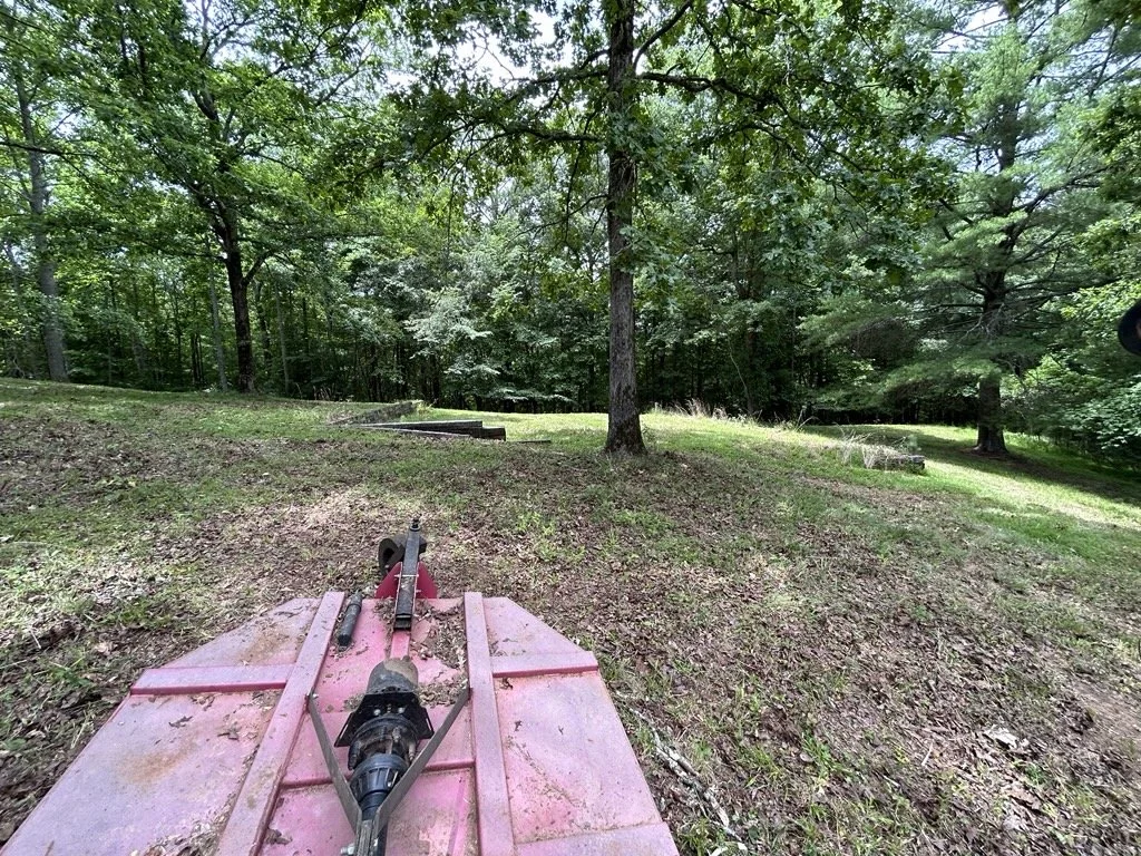 View from behind a pink mower on a grassy, wooded area with trees and a small hill.