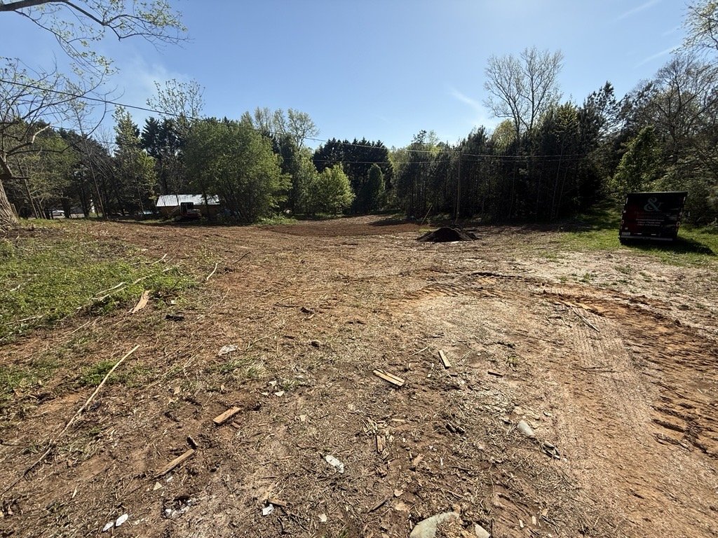 A cleared dirt lot with some scattered debris, surrounded by trees and a few parked vehicles under a clear blue sky.
