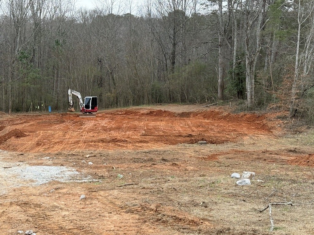 A construction site with orange dirt and a small excavator, surrounded by trees and a forest.