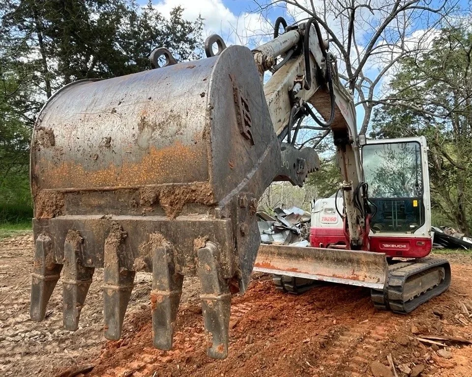 A small excavator with a bucket attachment at a construction or excavation site, surrounded by dirt and trees in the background.