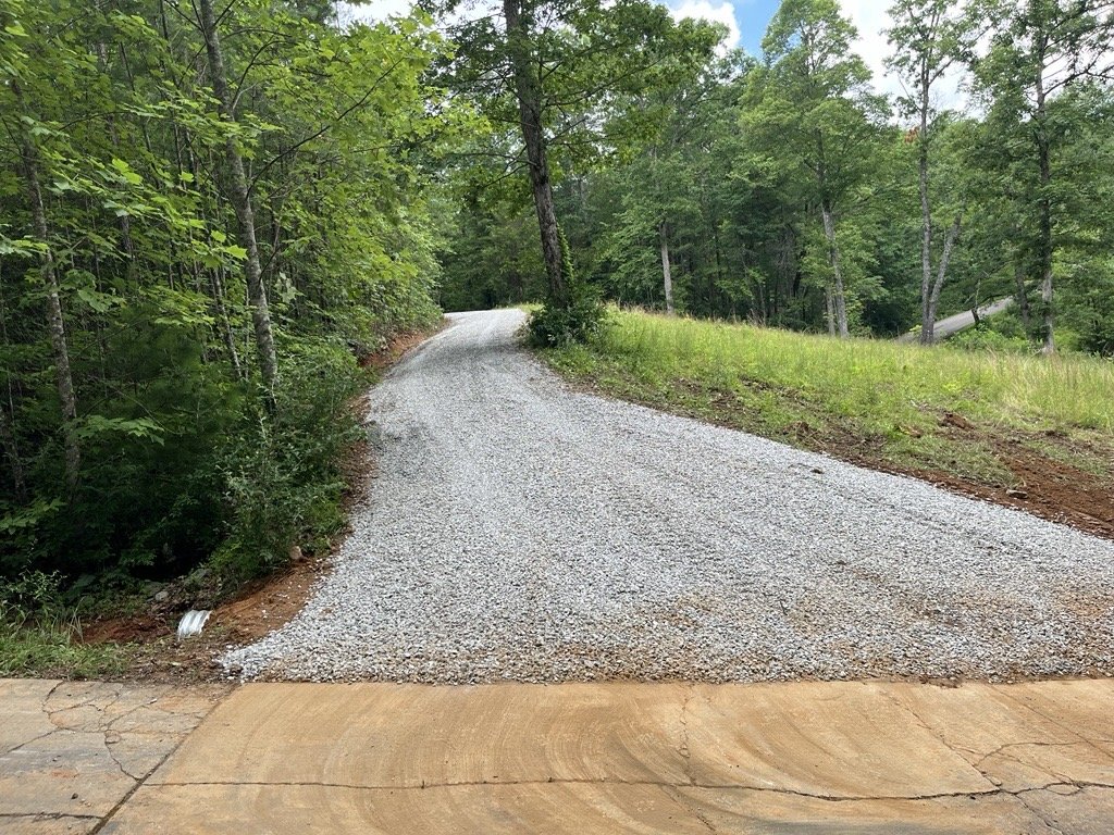 A gravel driveway leading into a wooded area with green trees and grass on either side.