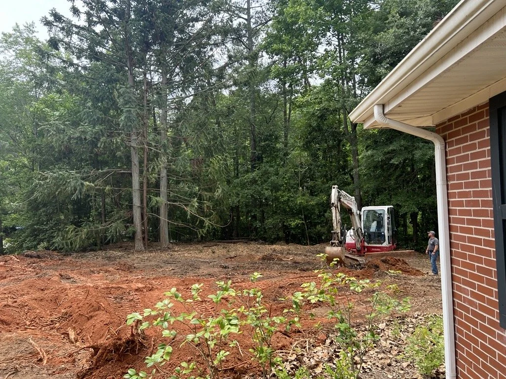 Construction site next to a brick house with a small excavator and a person, with a wooded area in the background.