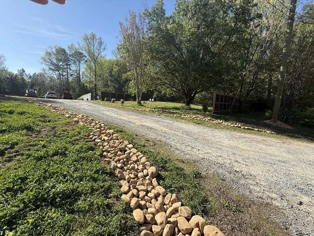 A gravel driveway bordered by a line of rounded rocks on the left side, with green grass and trees surrounding the area, under a clear blue sky.