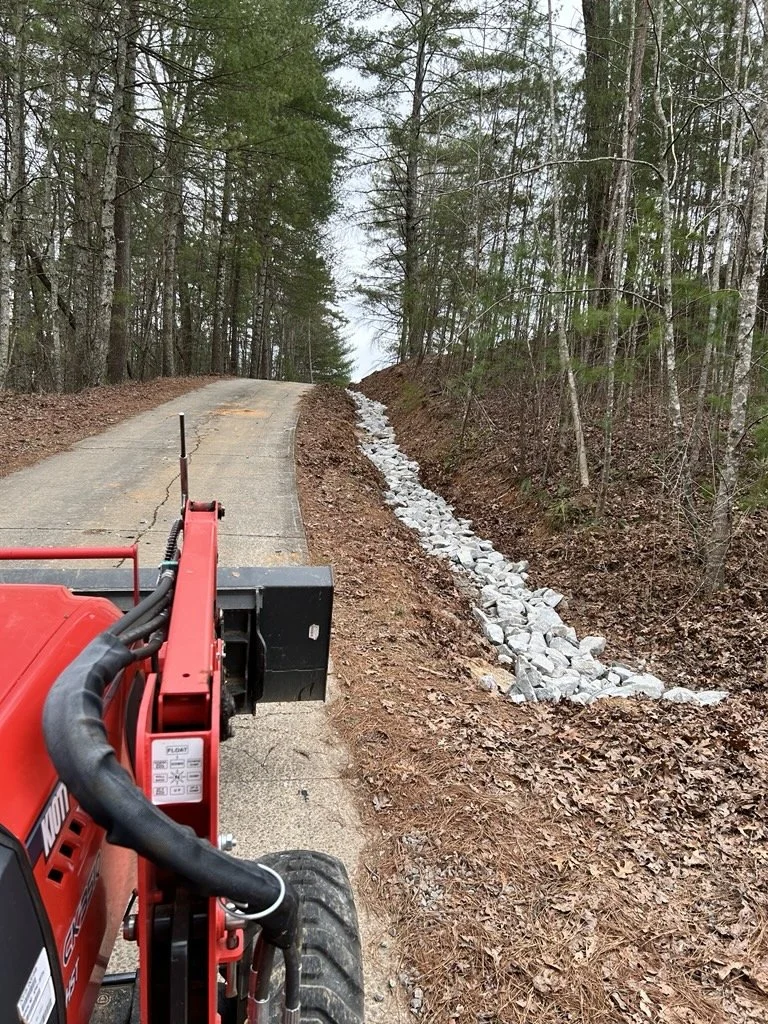 A small construction or landscaping machine on a dirt road next to a ditch filled with rocks, surrounded by trees.