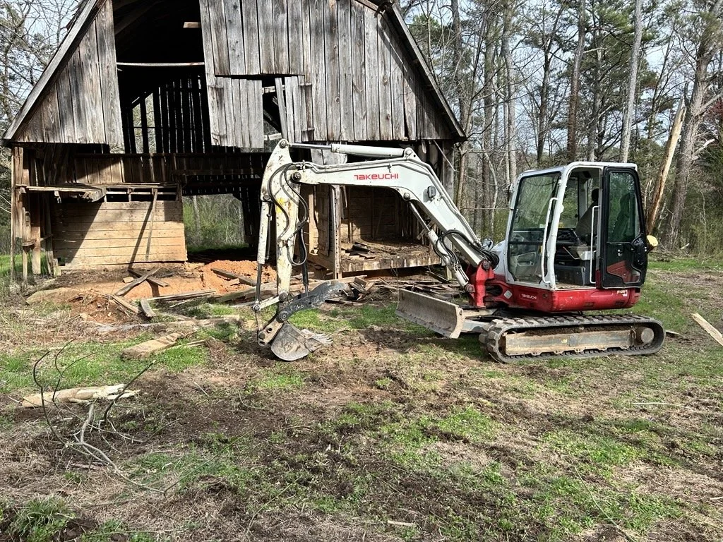 A small excavator demolishing a wooden barn in a rural area surrounded by trees with a partially cleared ground.