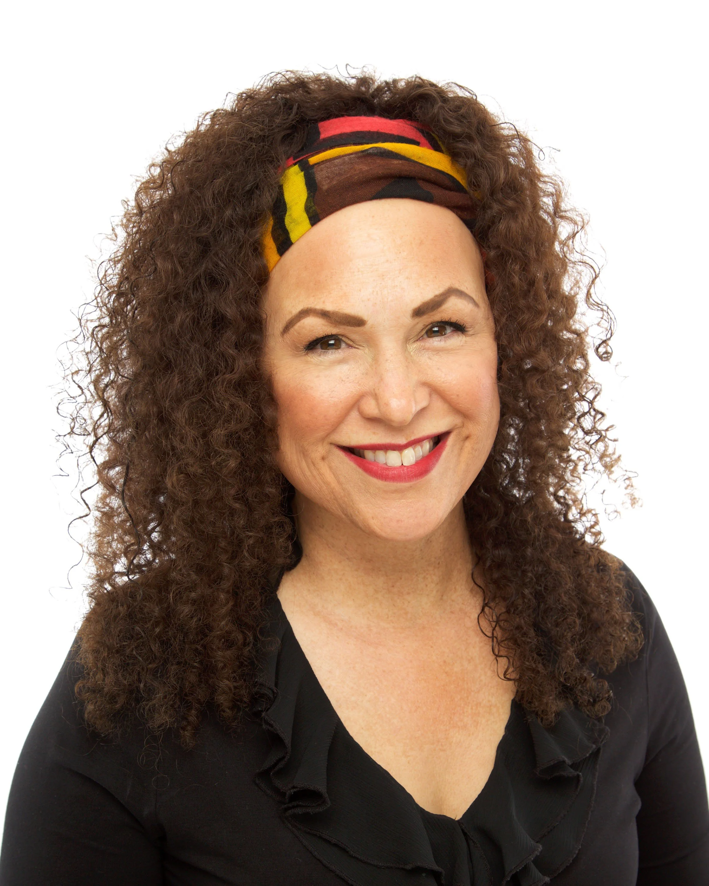 Close-up of a woman with curly brown hair, wearing a colorful headband and a black shirt with ruffled collar, smiling against a plain white background.