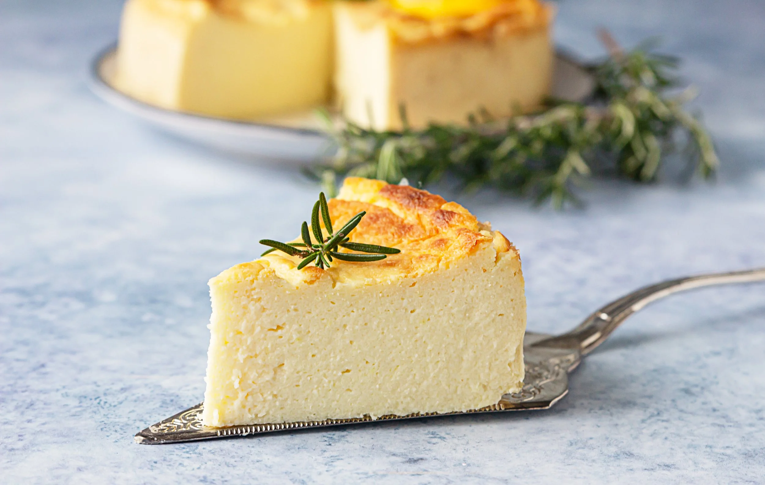 A slice of cheesecake garnished with a sprig of rosemary on a vintage silver dessert fork, with a whole cheesecake topped with sliced fruit and herbs in the background.