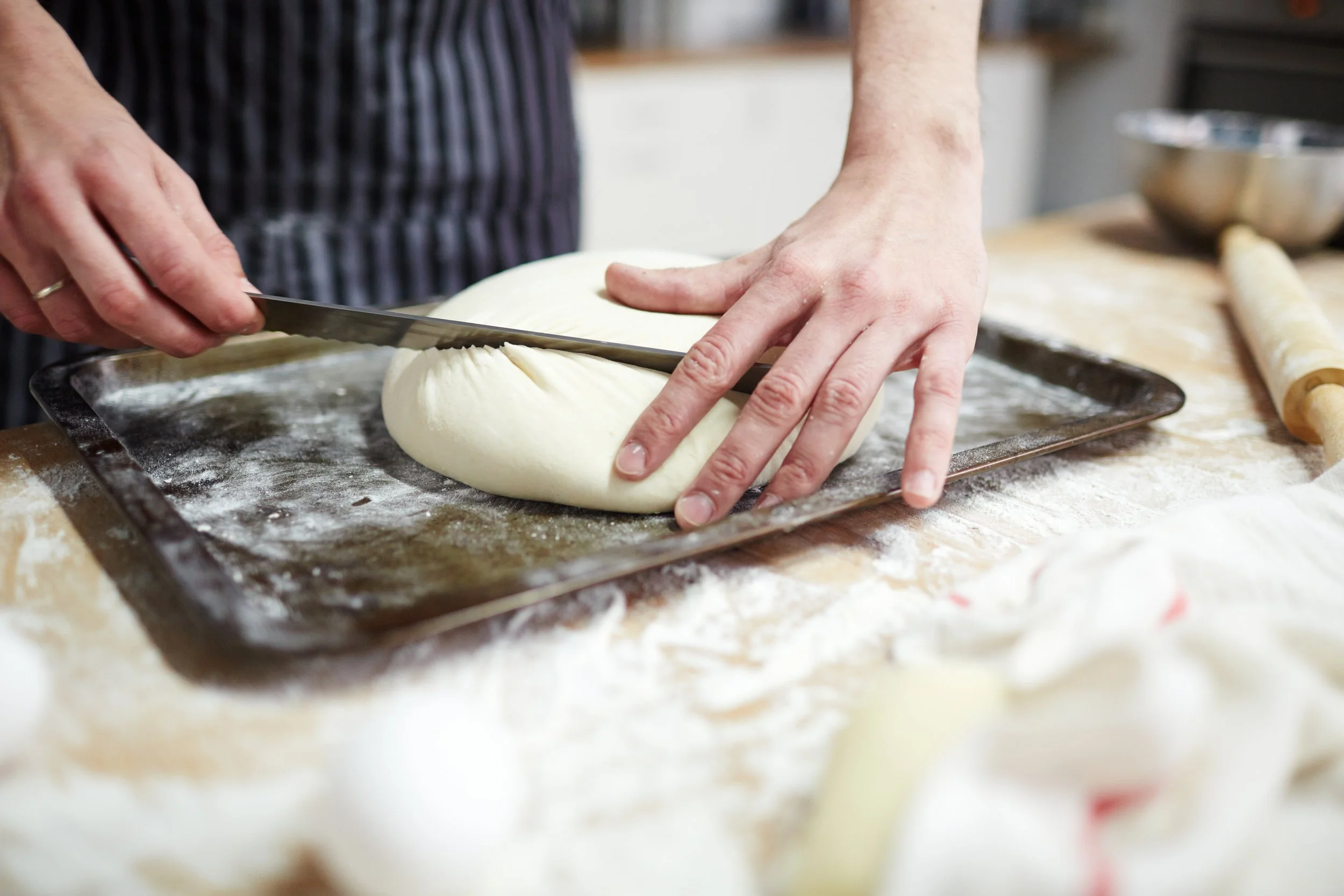 Person slicing dough on a baking sheet covered with flour in a kitchen.