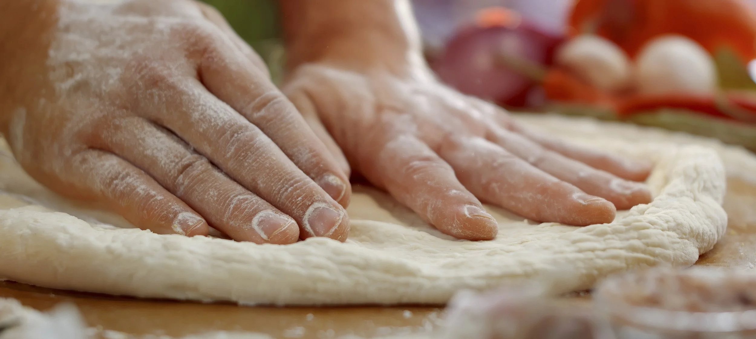 Hands dusted with flour pressing down dough on a surface, with tomatoes and eggs in the background.
