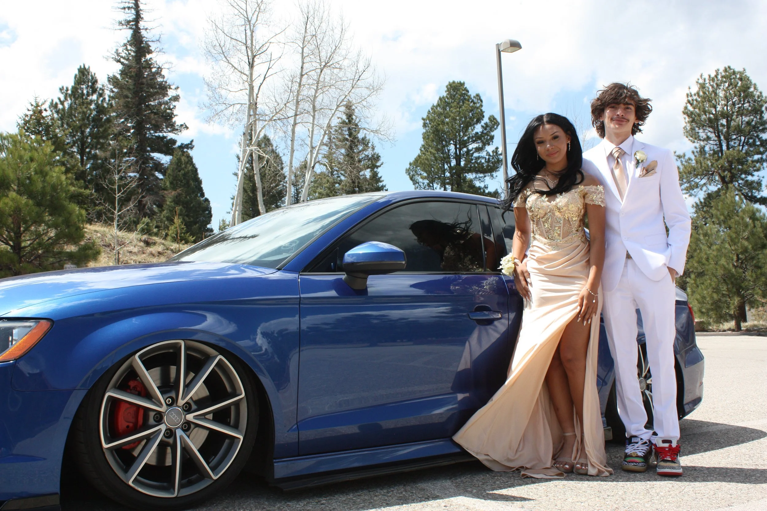 A young couple dressed in formal wedding attire standing next to a blue sports car outdoors with trees and a streetlamp in the background