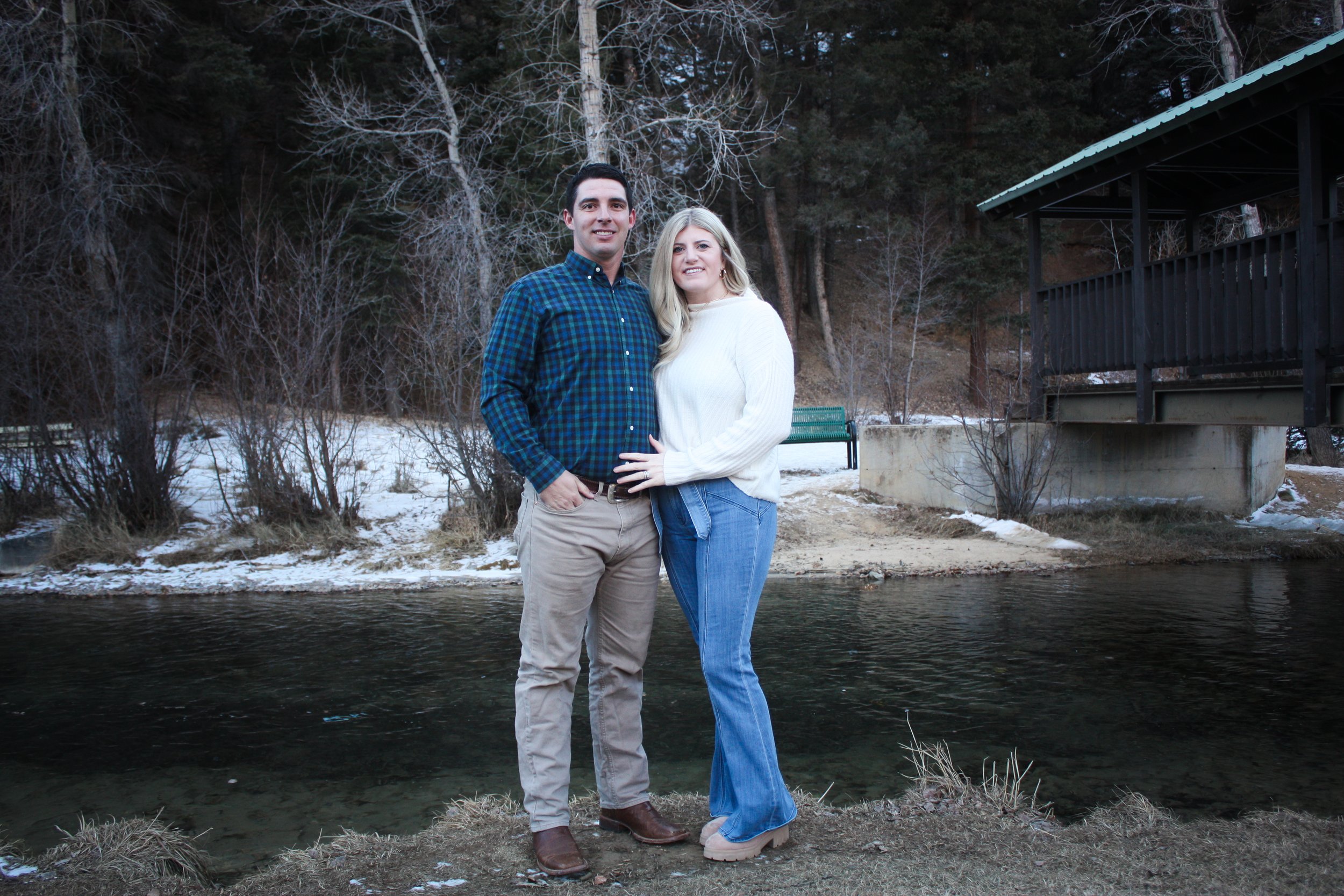 A couple standing by a river with a backdrop of leafless trees and a small constructed structure, possibly a bridge, on the right side of the image. It appears to be late fall or winter.