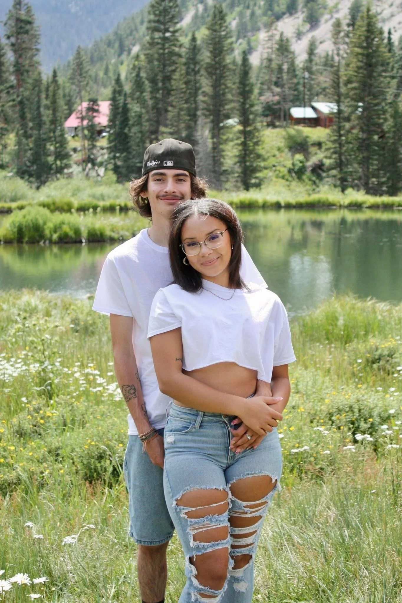 Two young adults, a man and a woman, standing close together outdoors in a lush green meadow with yellow and white flowers, by a calm lake with a forested mountain background, during daytime.