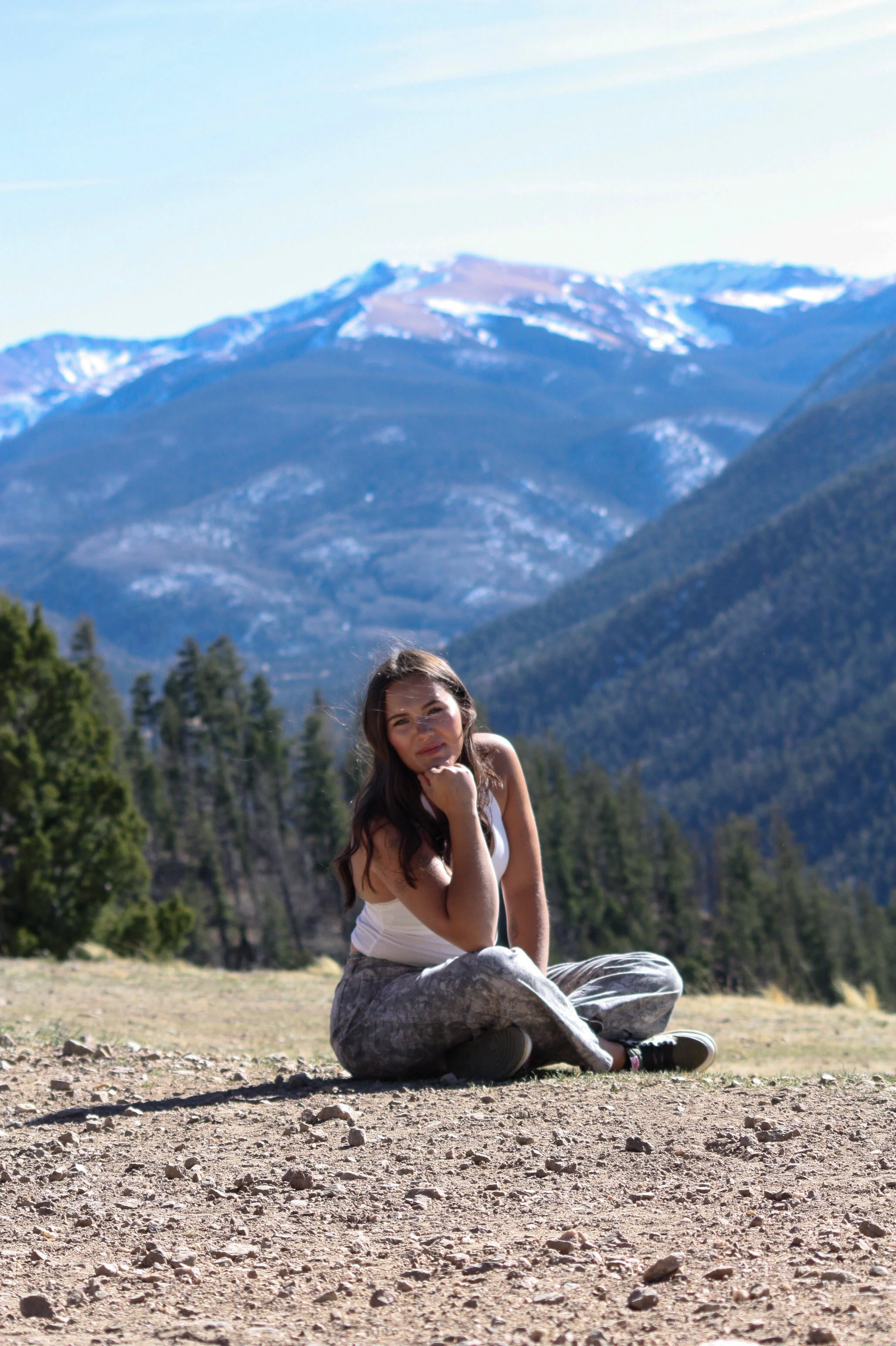 A woman sitting cross-legged on the ground in a mountain landscape, with snow-capped peaks and trees in the background, during daytime.