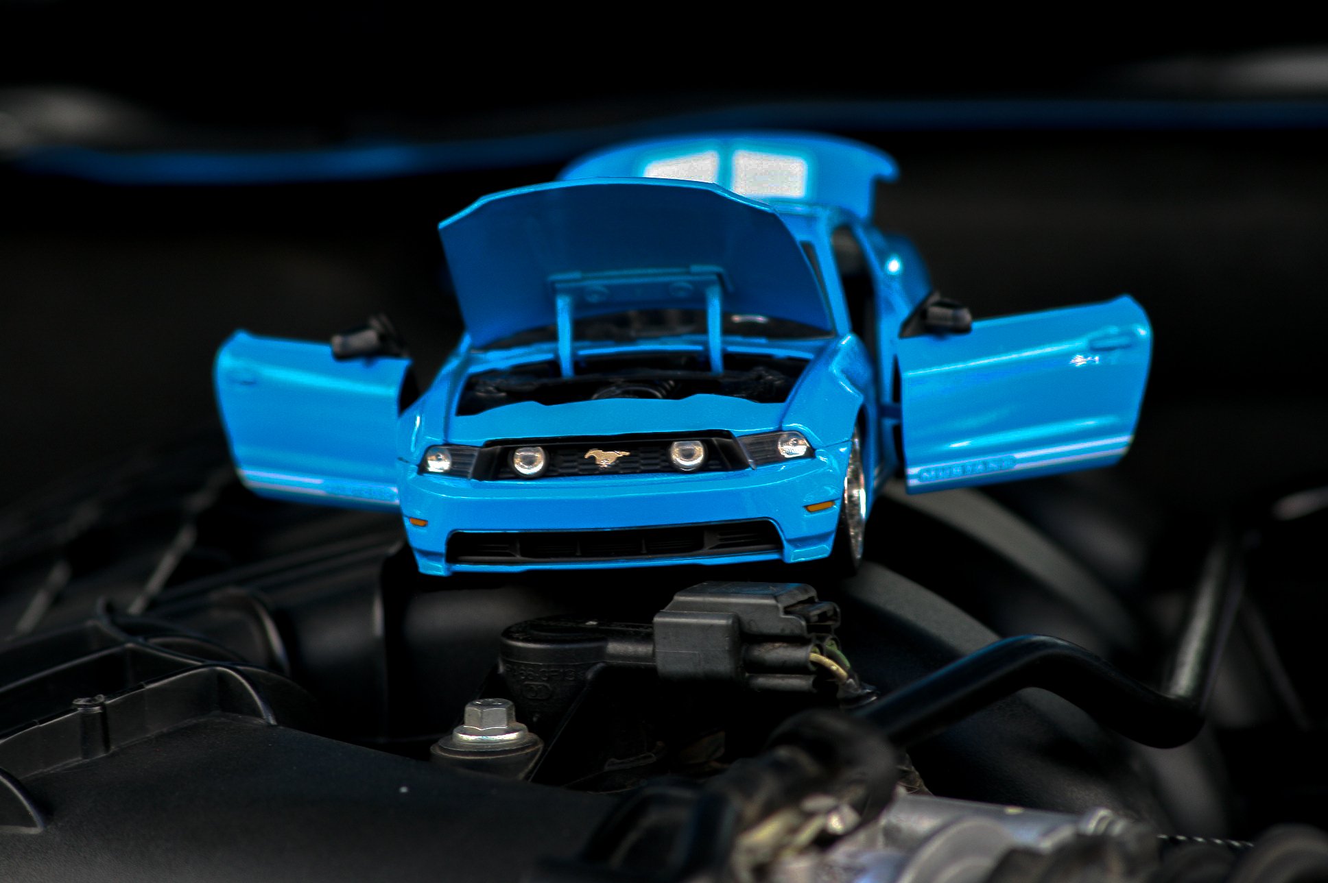 Small blue toy car with hood and doors open, placed on a black surface with wires and mechanical parts around.