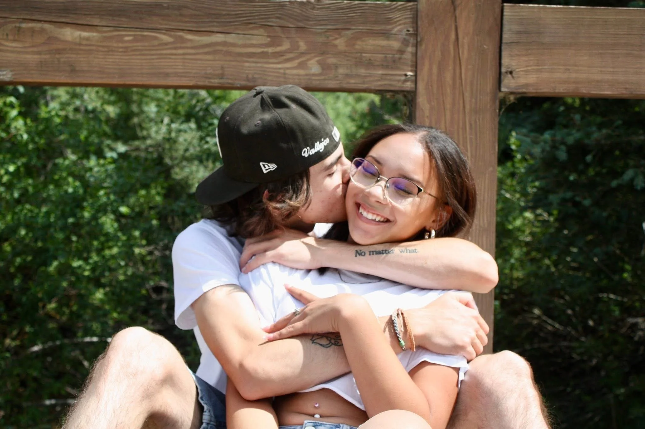 A young man and woman hugging outdoors, smiling with eyes closed. The man is wearing a black cap and white shirt, and the woman is wearing glasses and a white shirt with a tattoo on her arm. There are trees in the background.