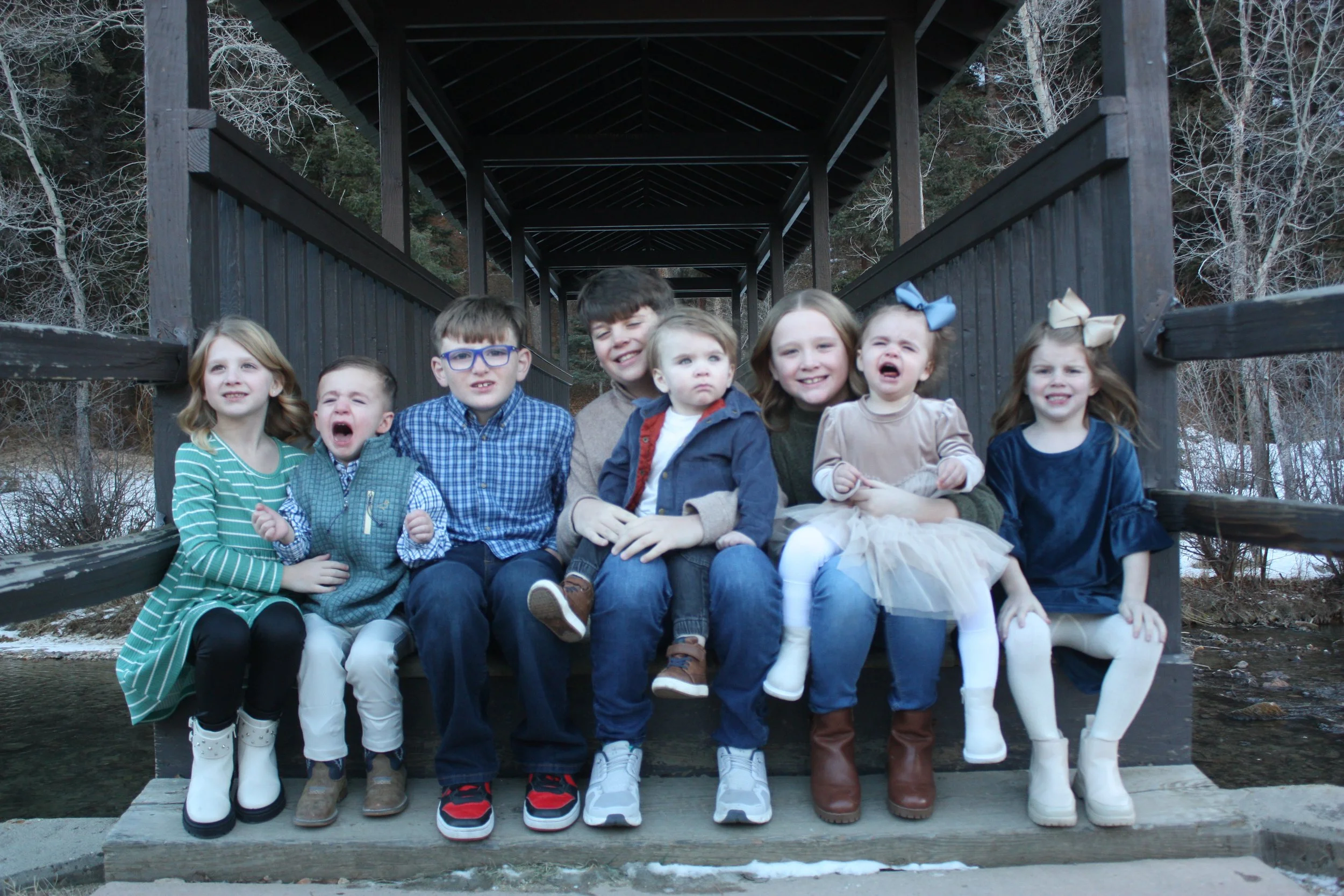 Group of children sitting on a wooden bridge outdoors, some smiling and others crying, with trees in the background.