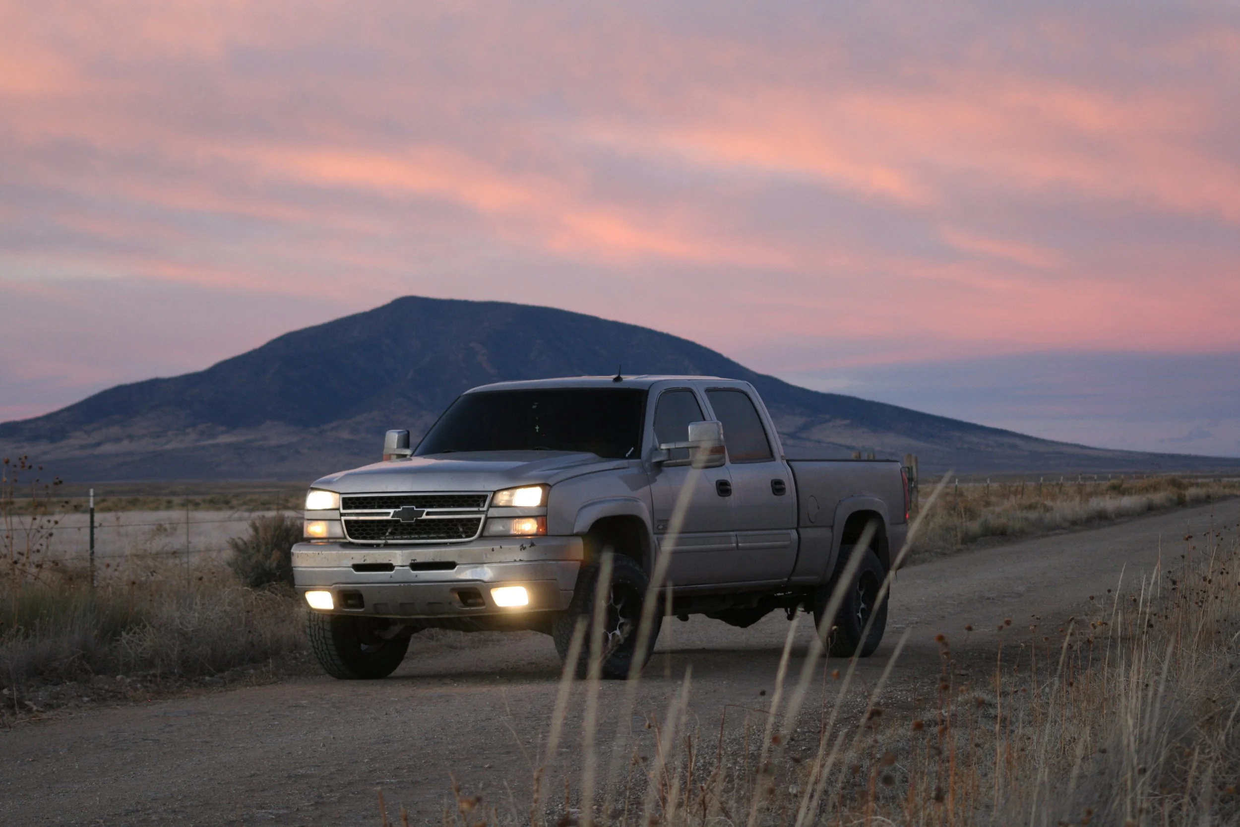 A silver Chevrolet pickup truck is driving on a dirt road in an open landscape during dusk, with a mountain and a pink and purple sky in the background.
