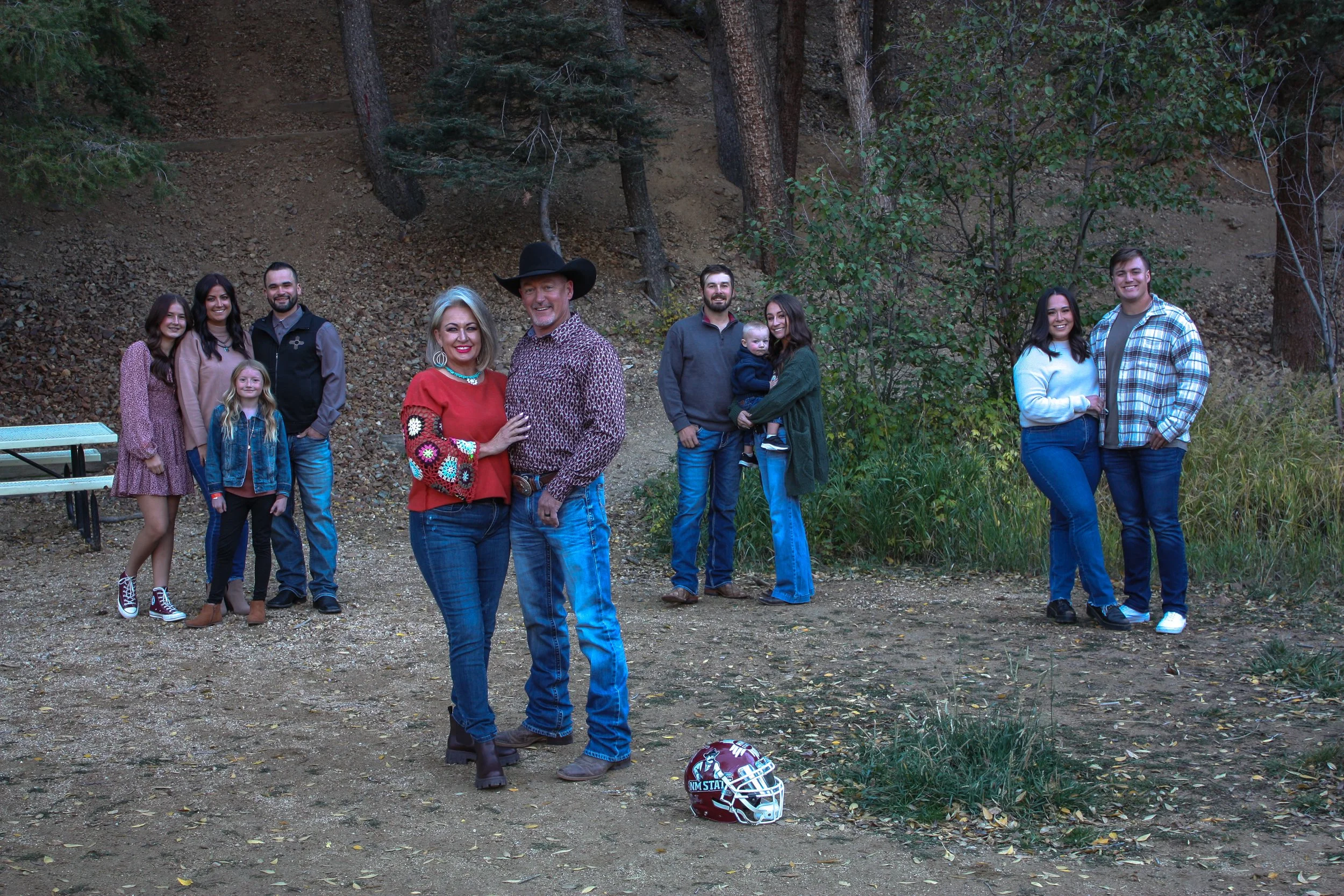 A large group of people, including several women, men, and children, standing outdoors in a wooded area with trees and foliage. Some are smiling, and there is a football helmet on the ground in the foreground.