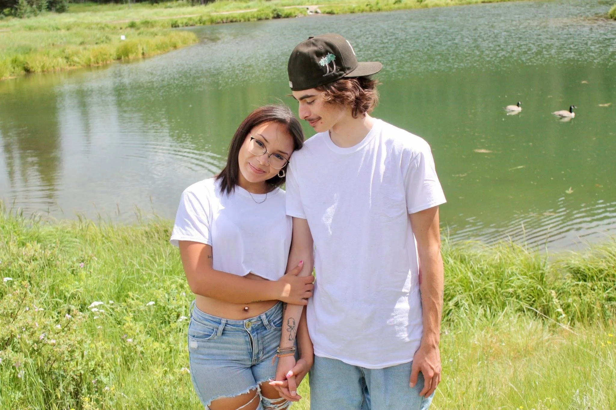 A young woman and man standing close together near a pond with geese, green grass, and trees in the background.