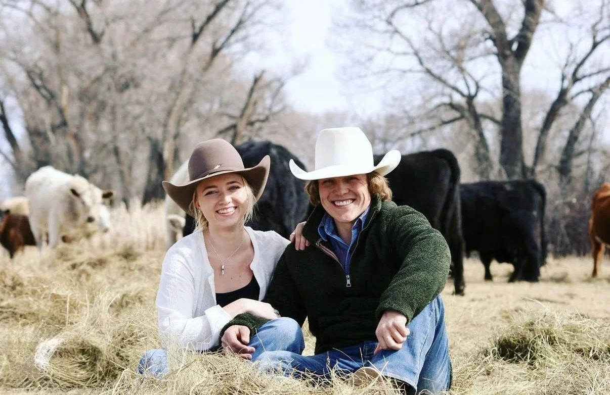 A smiling man and woman sitting on hay in a rural farm setting, wearing cowboy hats, with cows and trees in the background.