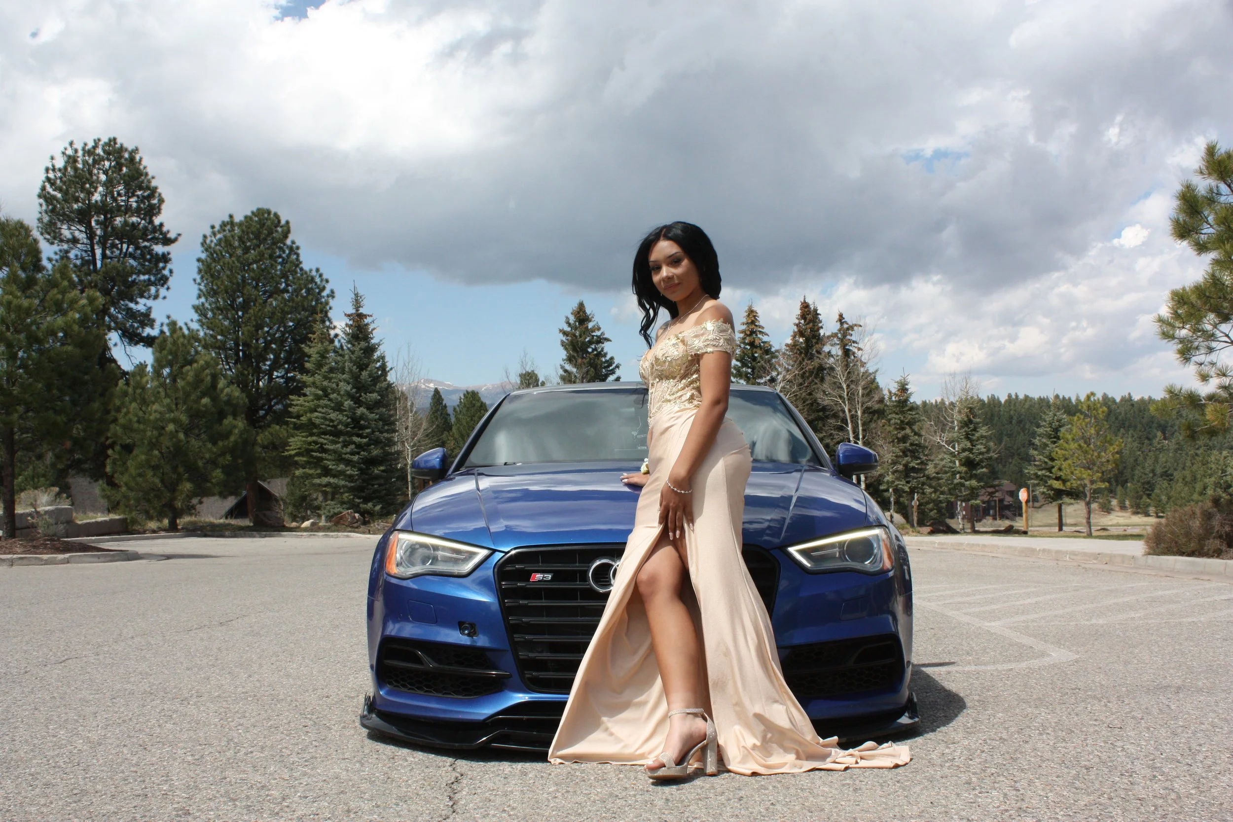A woman in a beige dress standing in front of a blue Audi car in an outdoor parking lot with trees and mountains in the background under a partly cloudy sky.