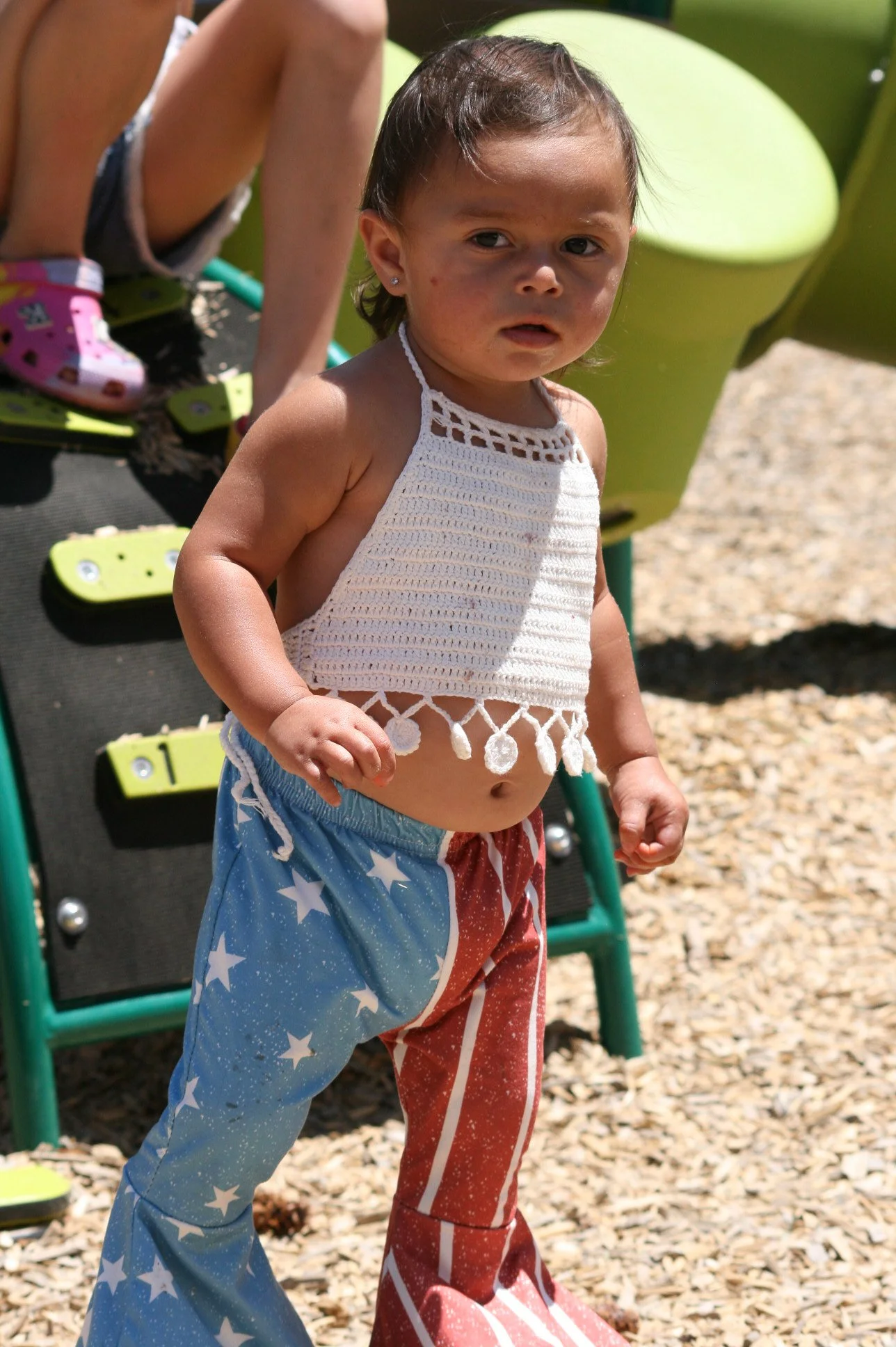 A young child with short dark hair and earrings, wearing a white crochet halter top and patriotic star-spangled pants, stands outdoors on a playground with wood chips on the ground, a slide in the background, and another child's legs visible.
