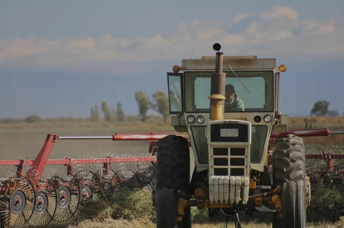 A tractor working in a field on a farm, with a person inside the cabin, under a partly cloudy sky.
