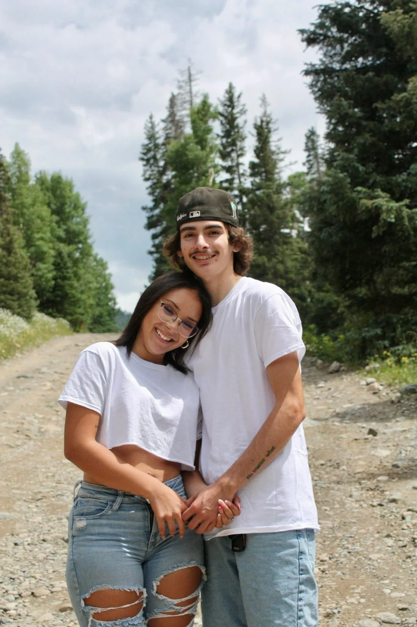 A young couple standing outdoors in a forested area, smiling and holding hands. The woman has long dark hair, wearing glasses, a white T-shirt, and ripped jeans. The man has wavy dark hair, a beard, wearing a backwards baseball cap, and a white T-shi