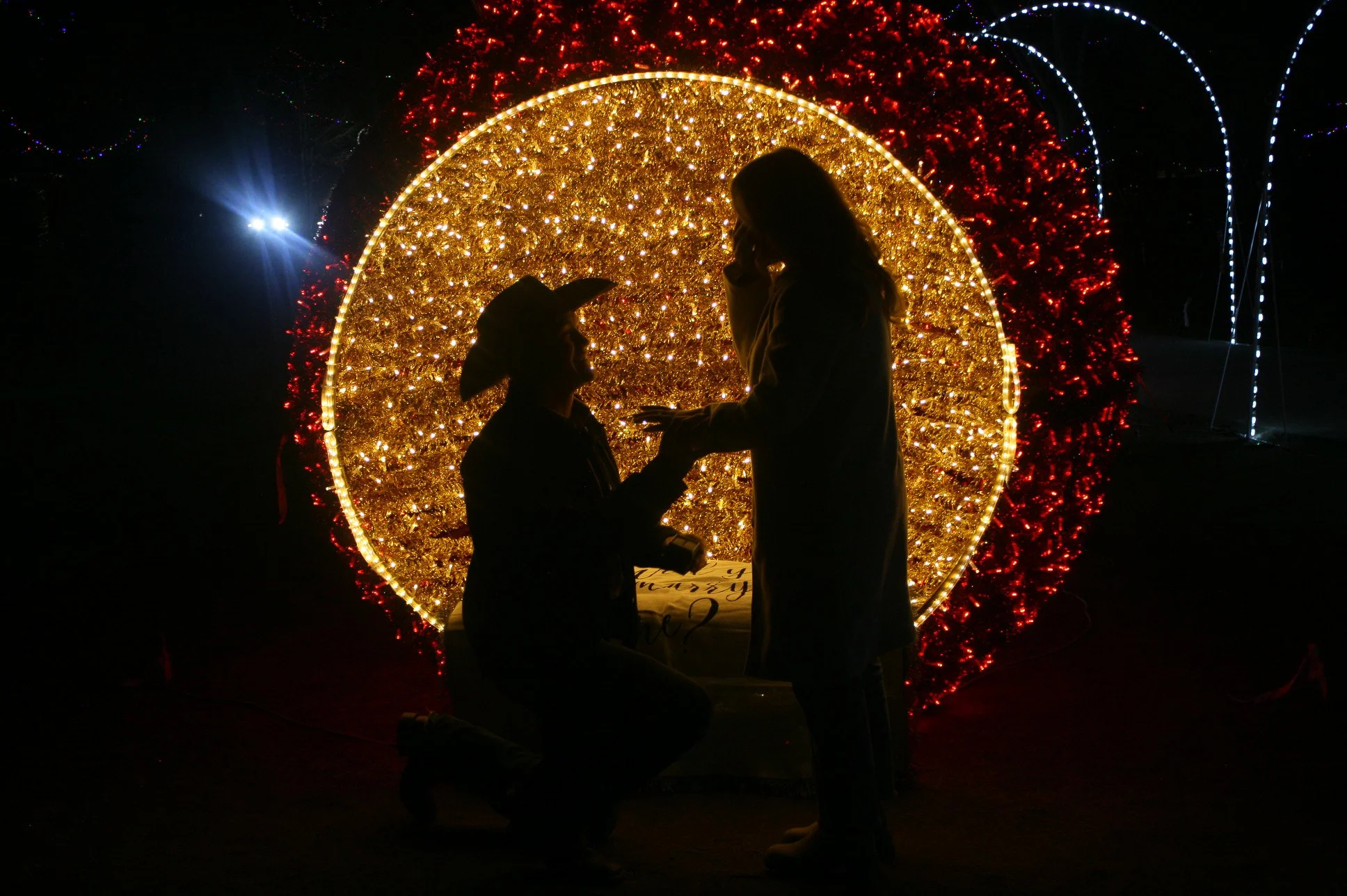 A couple is sharing a romantic moment during a proposal in front of a large, illuminated, gold and red holiday decoration at night, with the person kneeling and proposing to their partner.