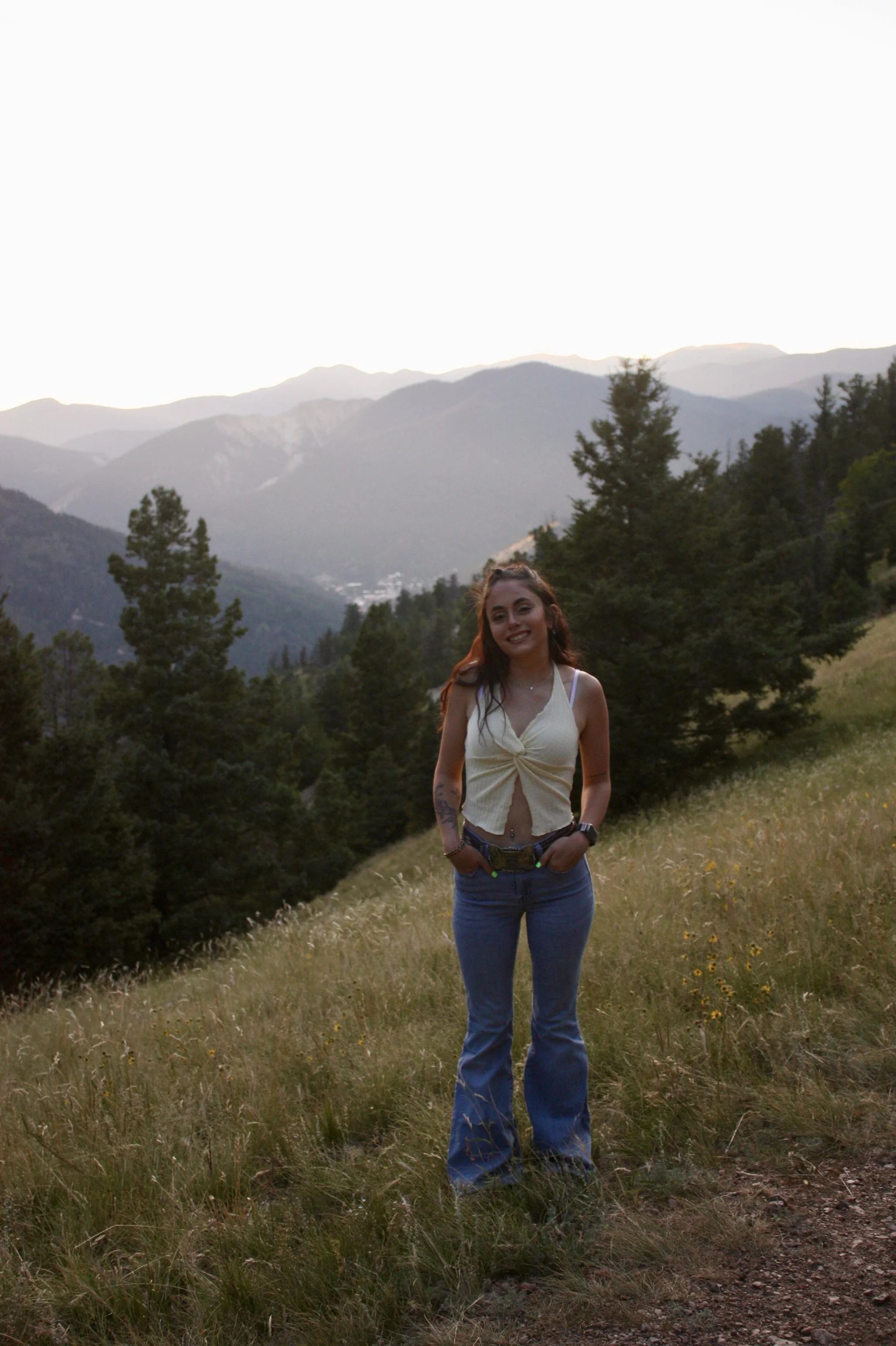 A woman with long reddish-brown hair, wearing a sleeveless pale yellow top and blue jeans, is standing in a grassy field with trees and mountains in the background during late afternoon or early evening.