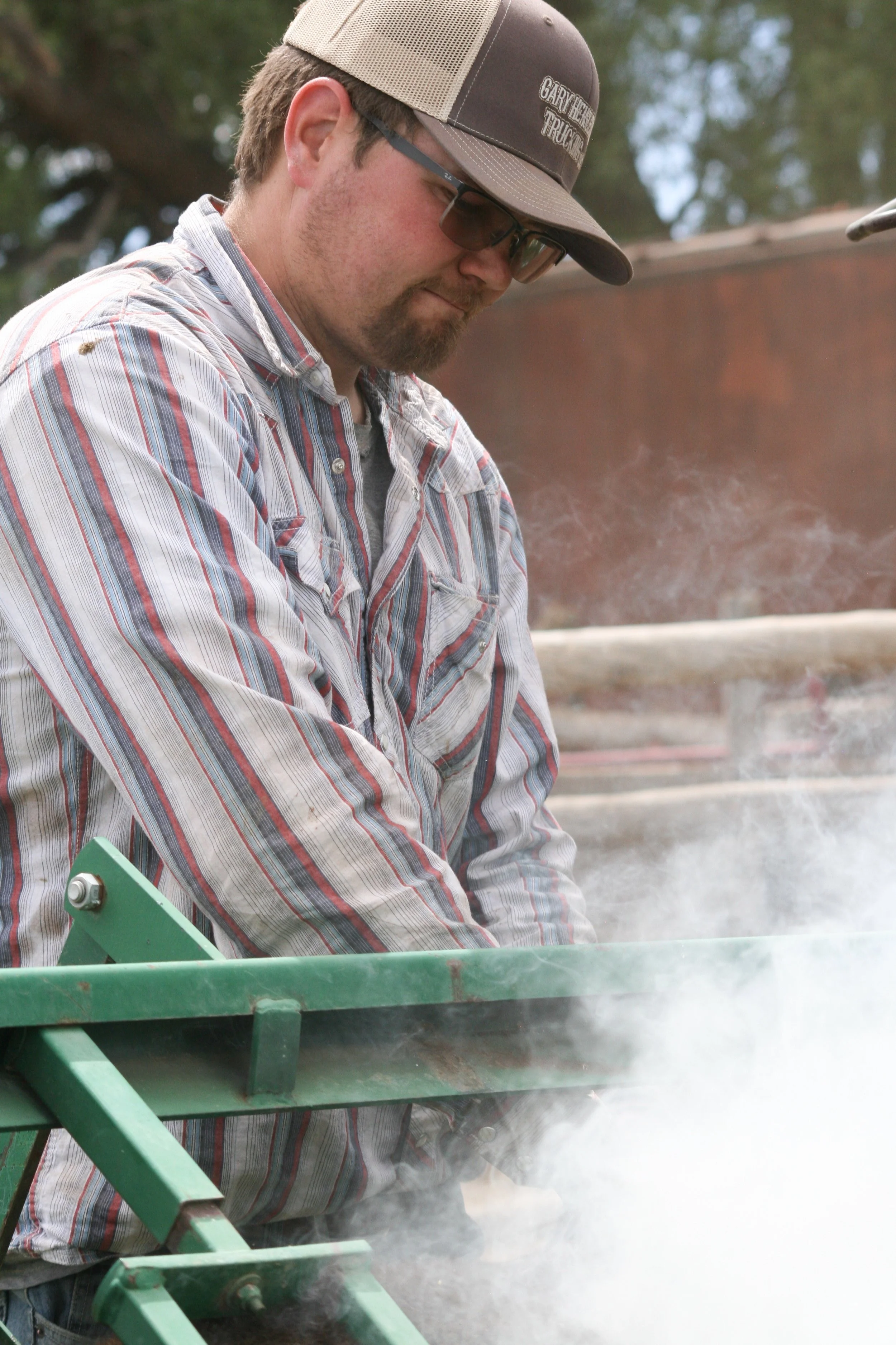 A man wearing a plaid shirt, sunglasses, and a trucker hat with the logo 'Garvin Trucking' is standing outdoors, looking down, near a green metal structure with wood and dirt in the background.