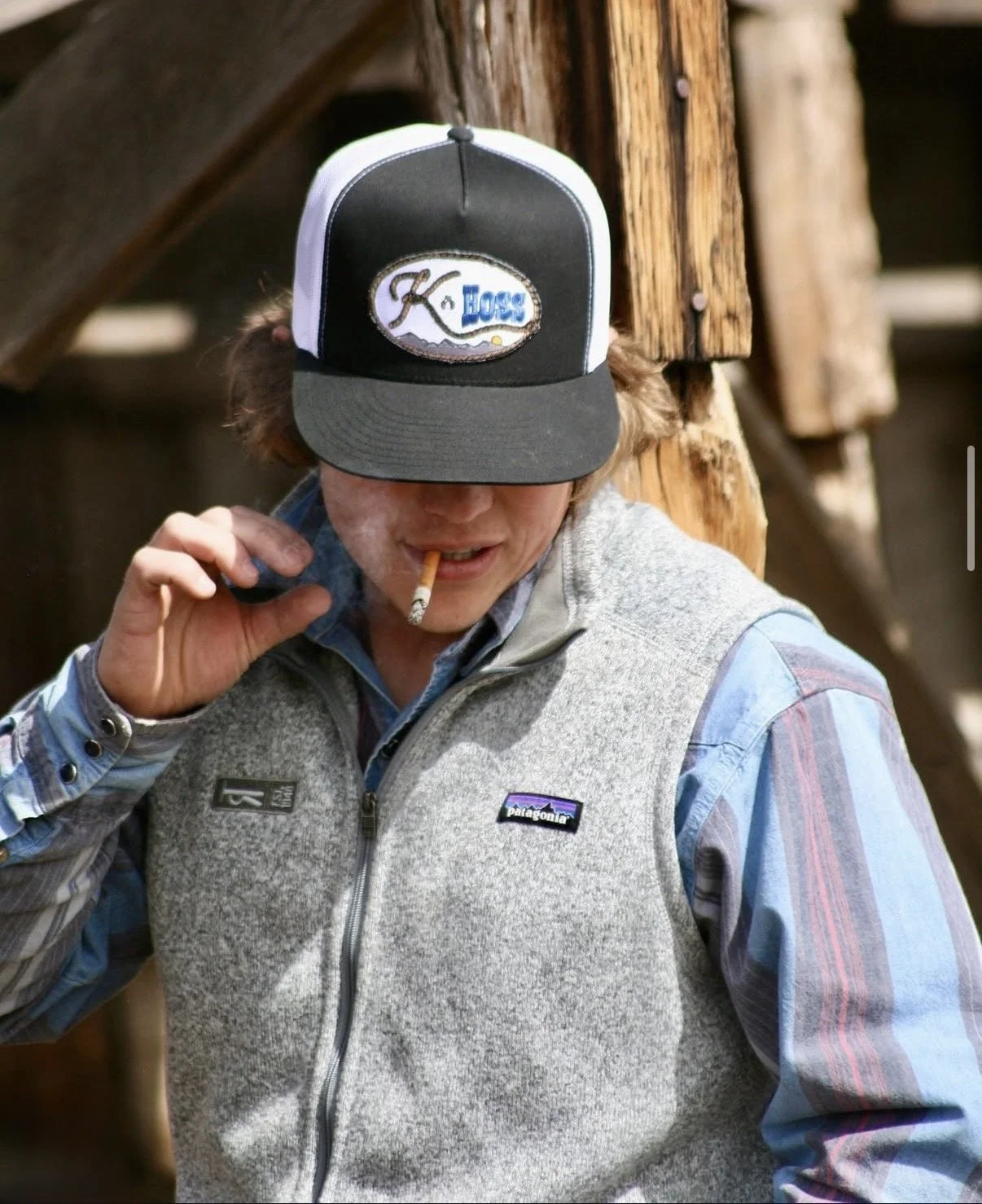 A man with a cigarette in his mouth, wearing a Patagonia vest and a black, white, and gray trucker hat with a logo that says 'K for Hogs', standing outdoors near a wooden structure.