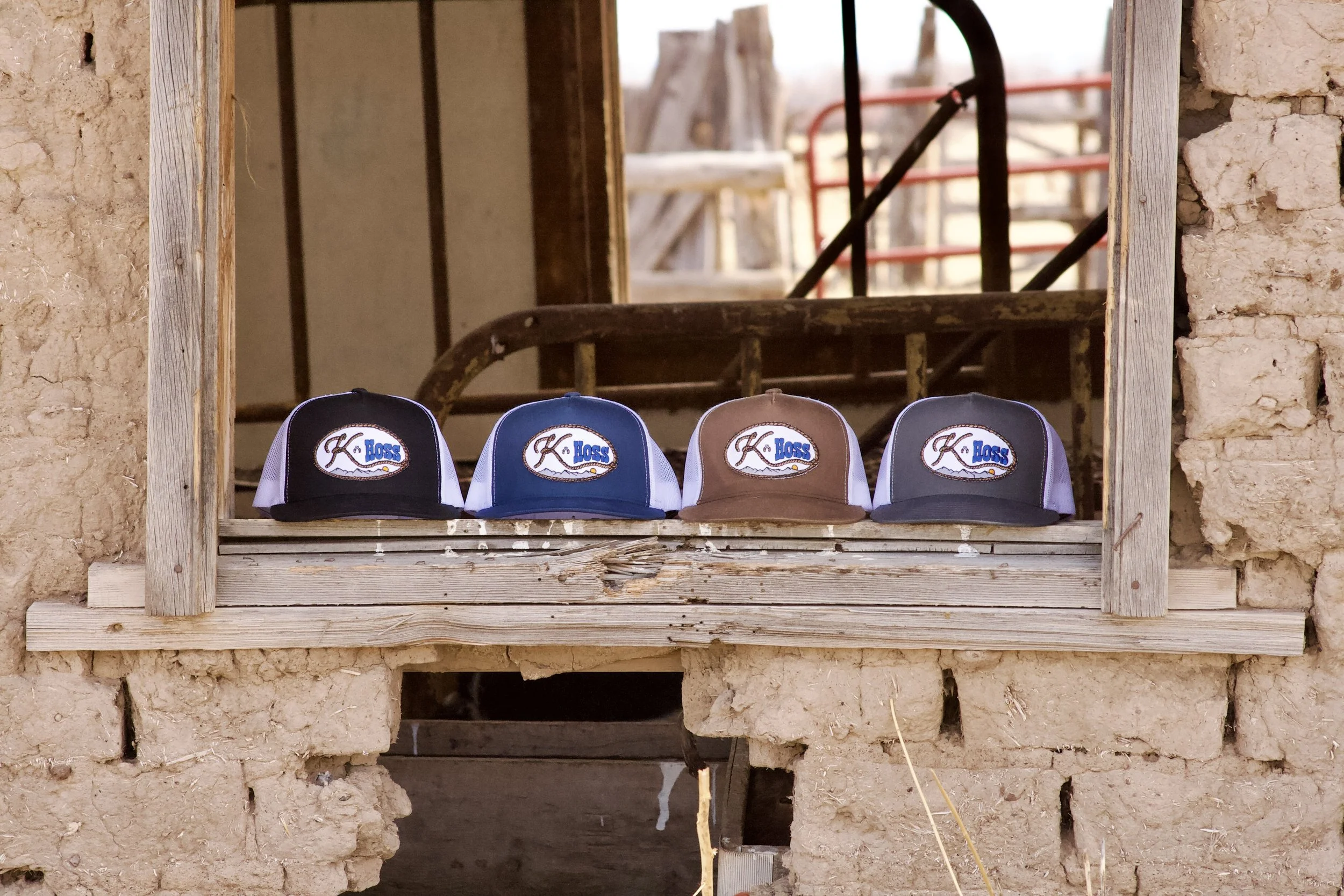 Four baseball caps with the logo 'K. Hoss' displayed on a wooden shelf in front of a rustic unfinished wall. The background shows construction tools and materials.