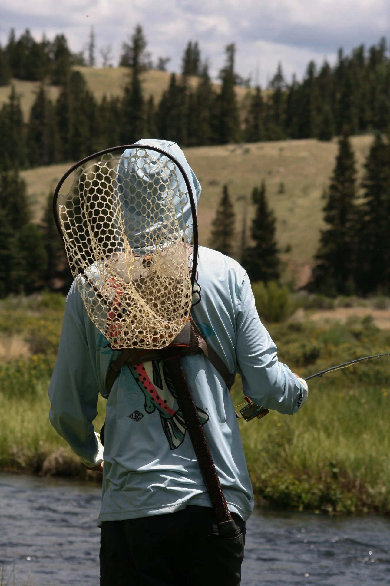 Person dressed in outdoor clothing holding a fishing rod, with a net hanging from their back, standing by a river in a wooded, hilly landscape.