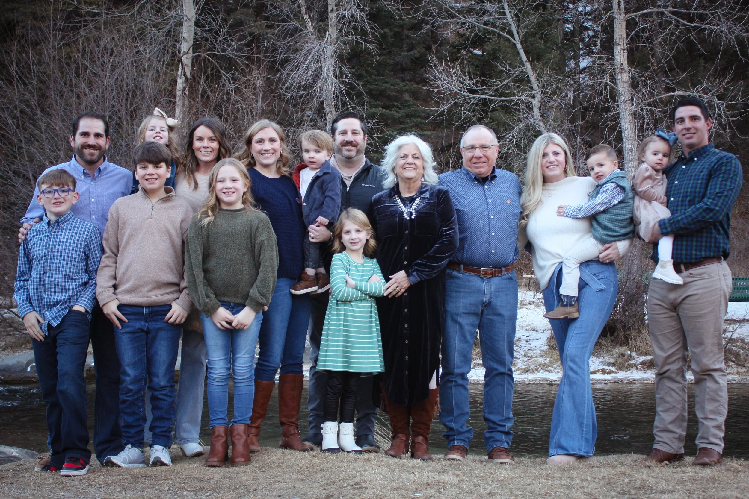 A large multigenerational family group outdoors in a wooded area with some snow on the ground. The family members are standing close together, smiling at the camera, with trees and a river in the background.