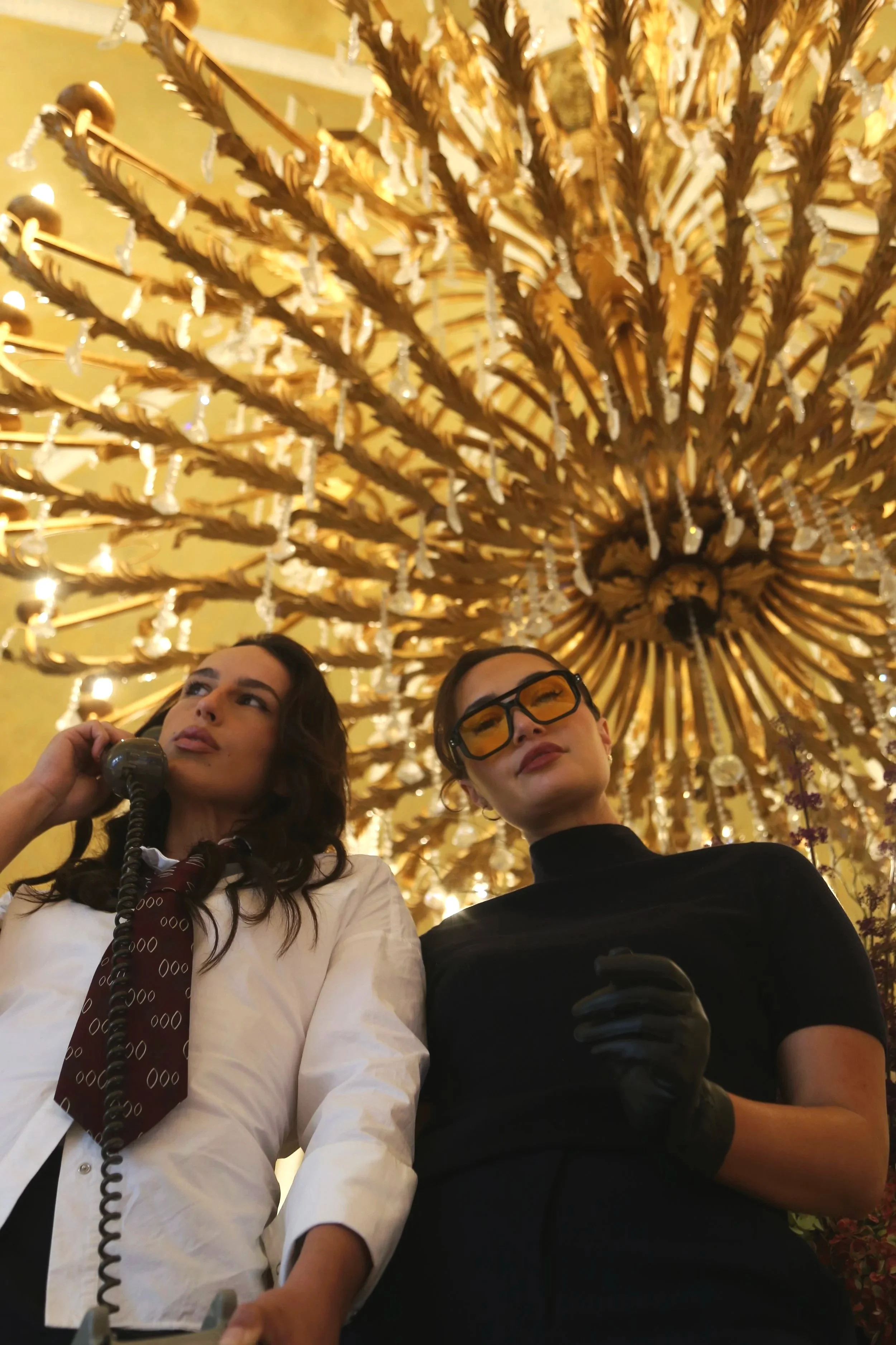 Two women are in a luxurious interior with a large ornate golden chandelier above. One woman is wearing a white shirt and a brown tie with a pattern, and is speaking into a corded phone. The other woman, wearing glasses and a black top, is looking up slightly. The scene suggests they are in a formal or elegant setting.
