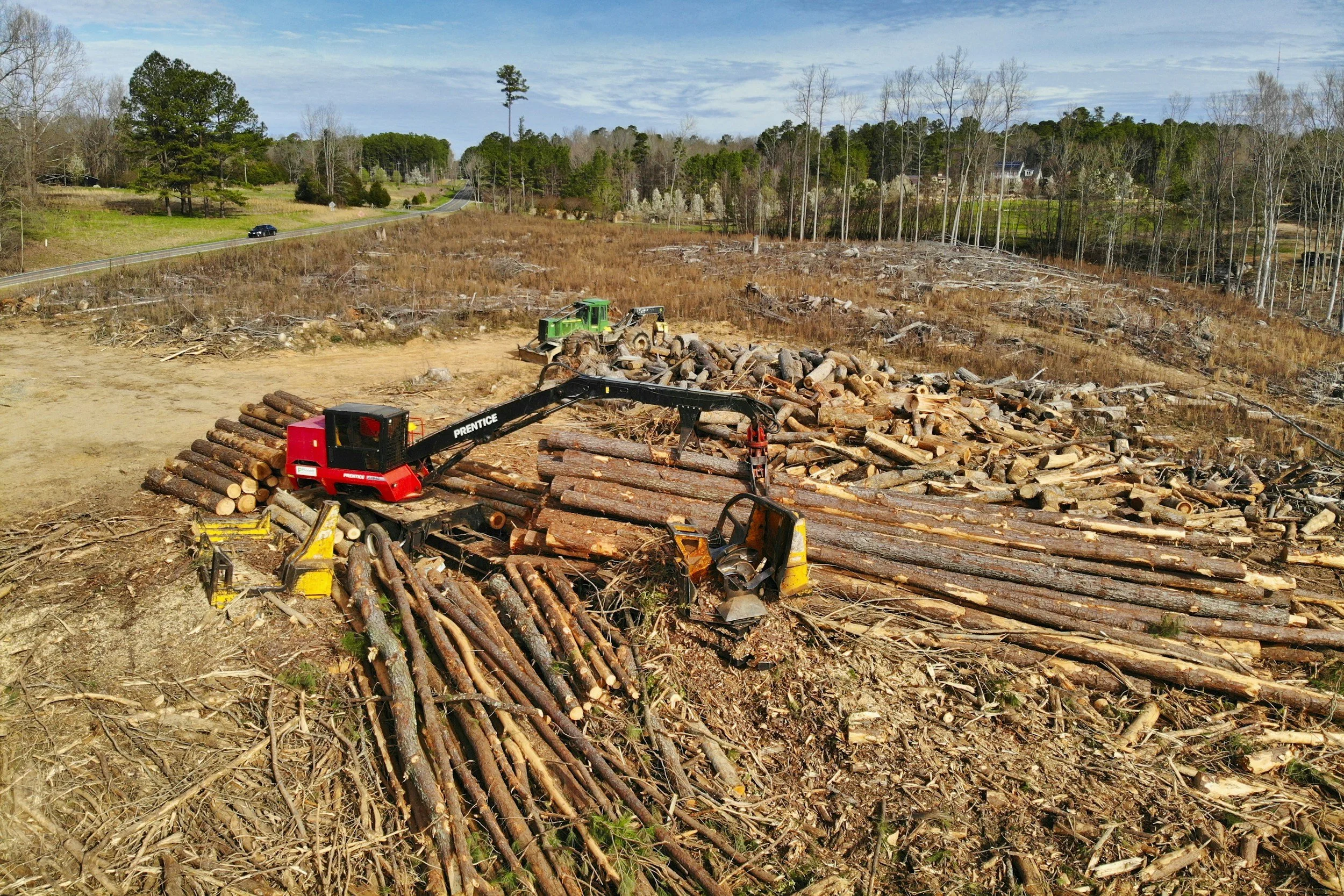 A land clearing scene with a pile of logs and wood debris, a green forestry machine, and a rural landscape with trees and a road in the background.