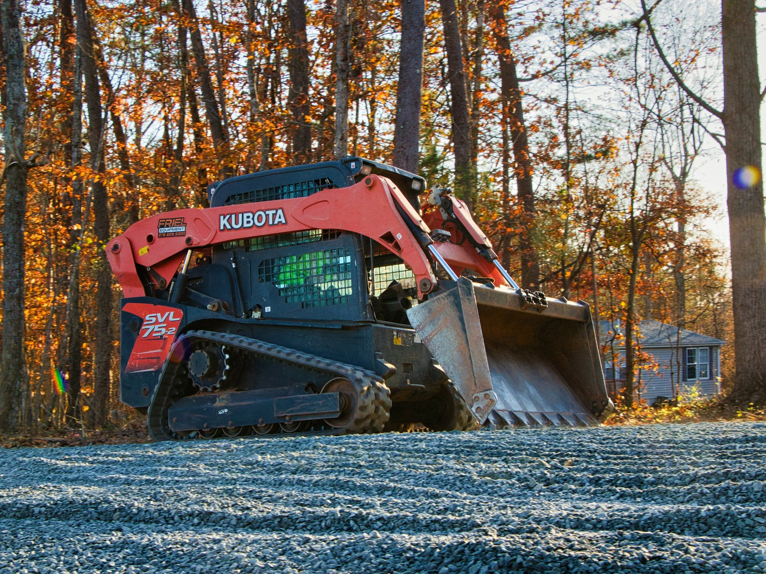 A red Kubota compact utility loader with a scoop attachment, operating on a gravel surface in a wooded area during autumn, with trees having orange and yellow leaves and houses visible in the background.