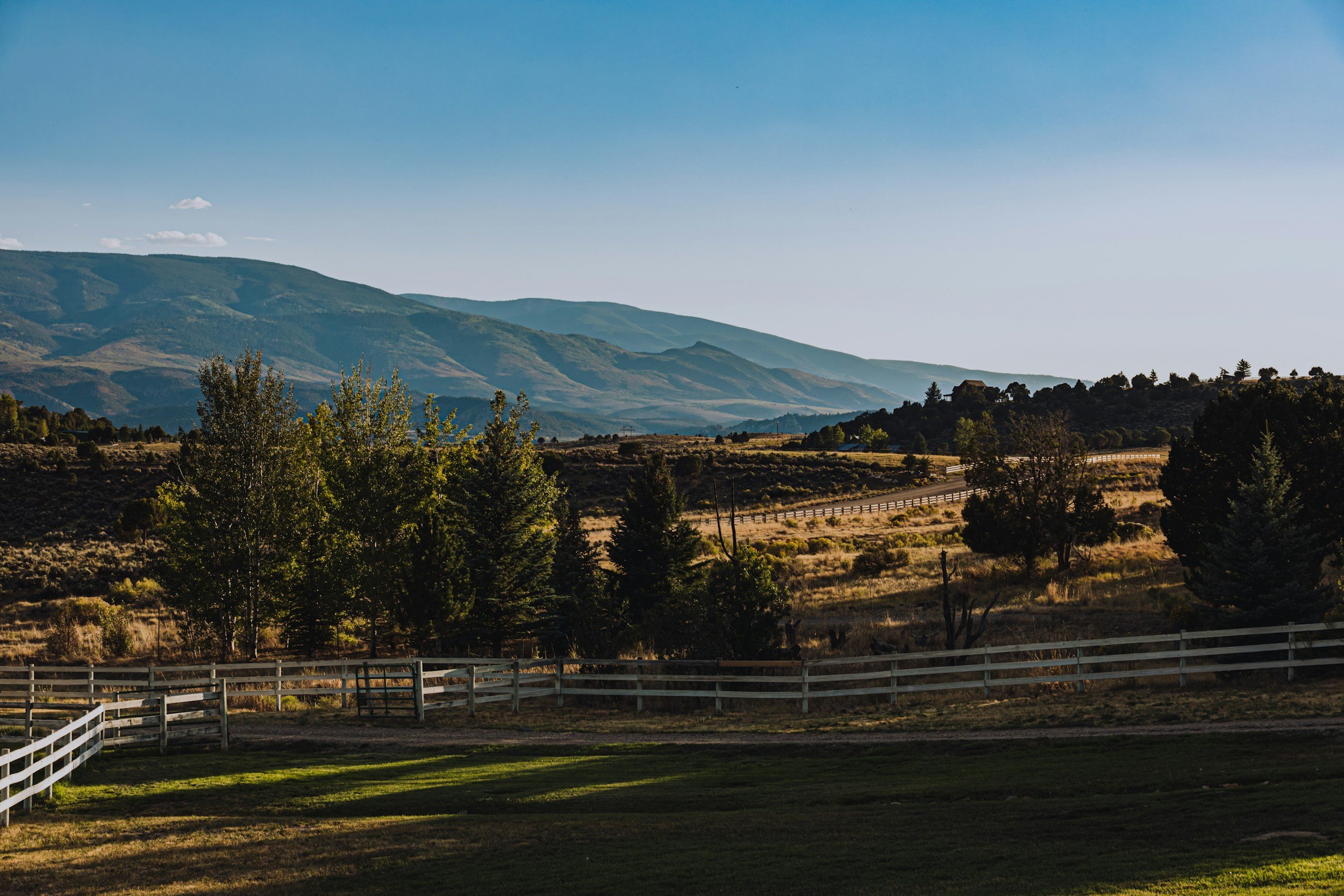 Scenic view of rolling hills and mountains under a clear blue sky with some sparse clouds, seen from a fenced pasture area with trees and grass in the foreground.