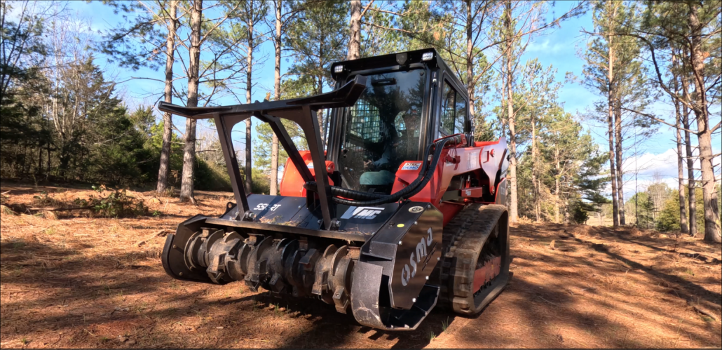 A compact red and black tracked skid steer loader on a cleared outdoor construction site with dirt and trees.