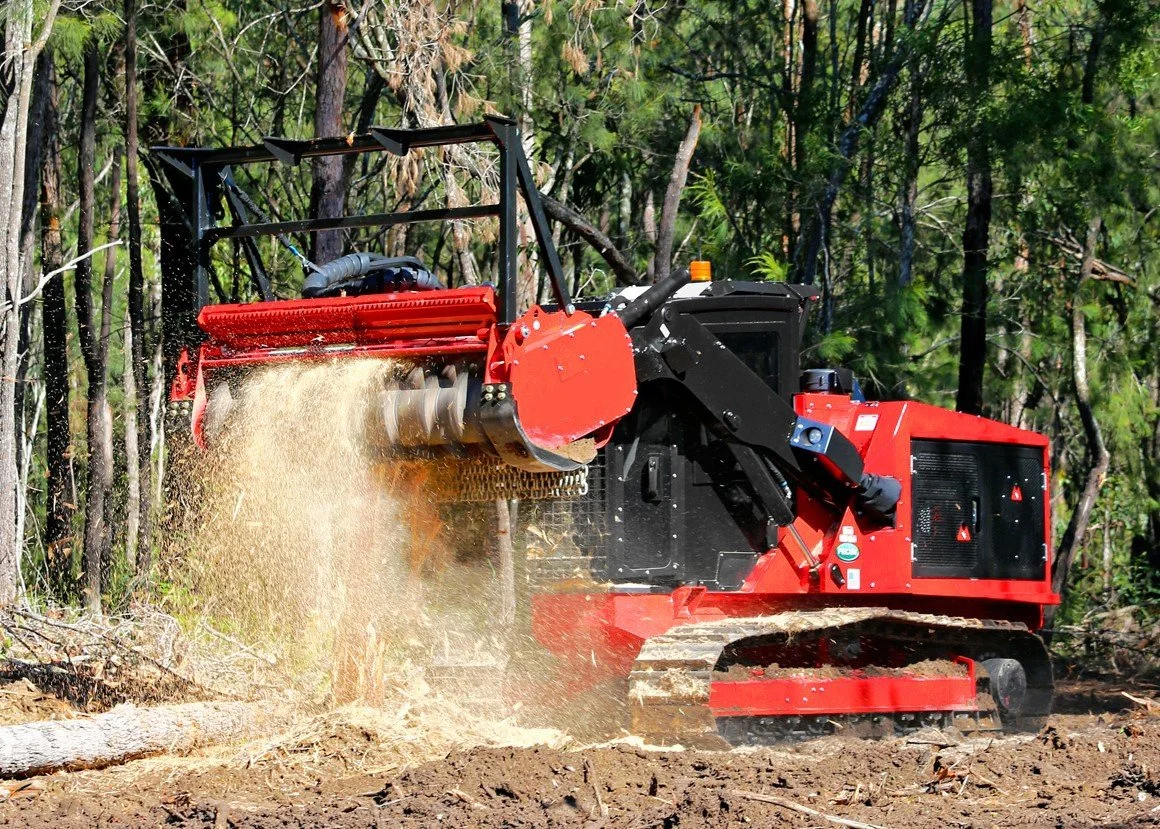 A red and black forestry mulcher attachment on a tracked machine clearing a forested area.