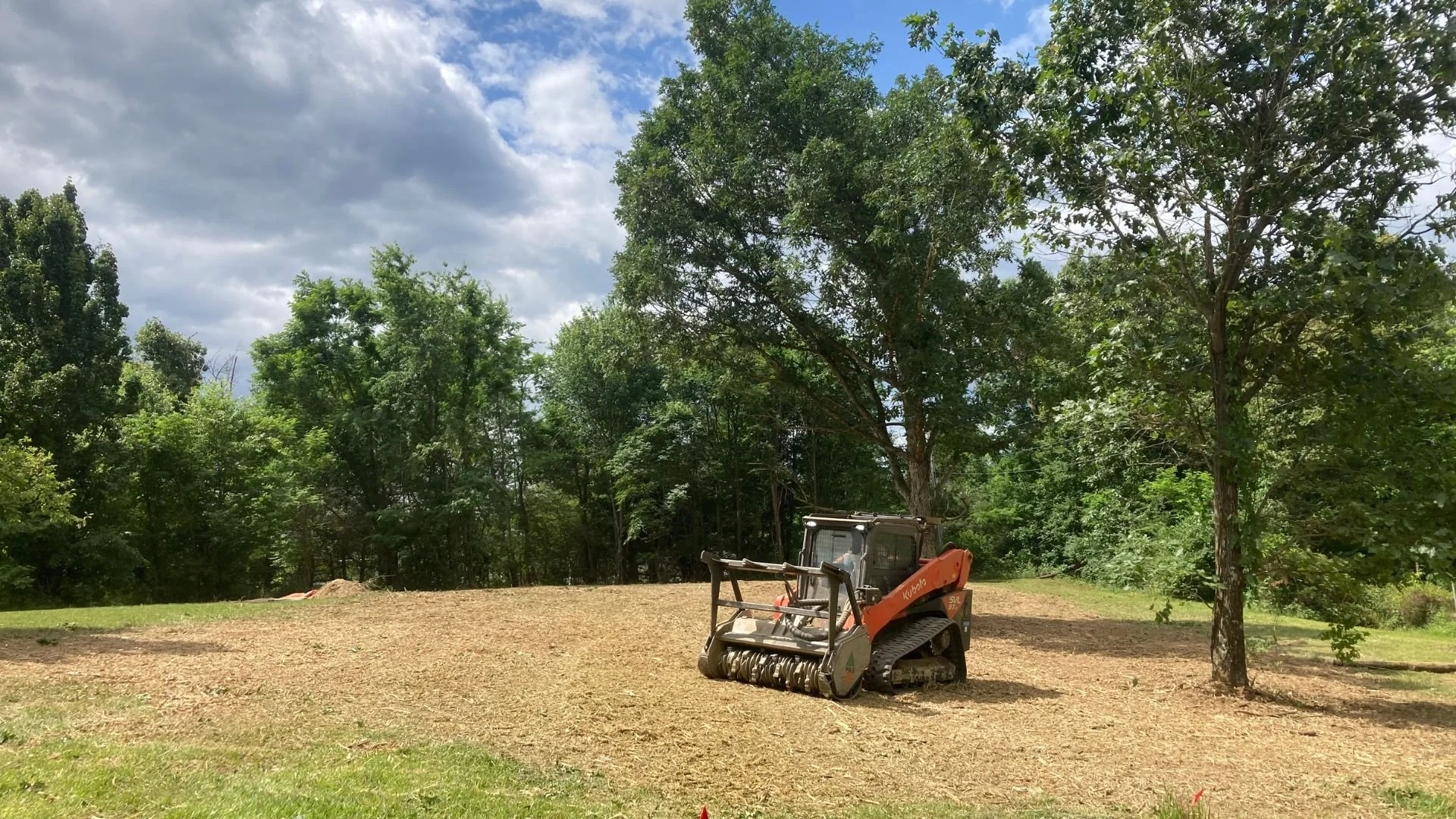A small orange Kubota mini skid steer with a black attachment on a cleared, dirt-covered area surrounded by green trees under a partly cloudy sky.
