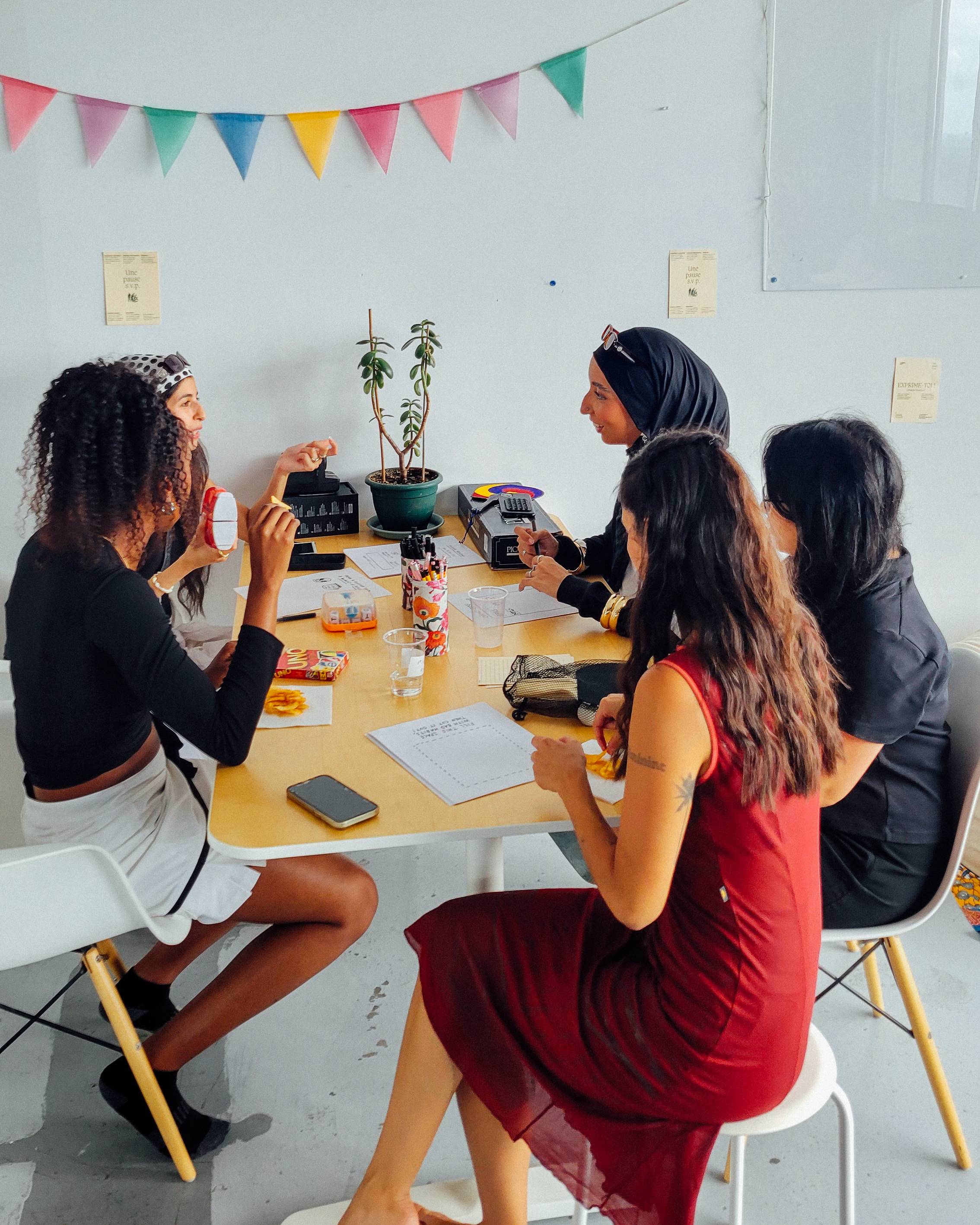 Groupe de femmes assises autour d'une table en train de discuter lors d'une fête ou d'une réunion informelle, avec une décoration de fanions colorés suspendus au plafond.